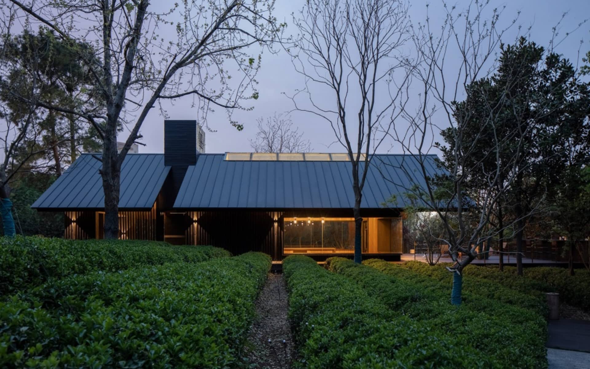 A modern house with a black roof and a green lawn, surrounded by trees, featuring a large window for natural light and a clean, minimalistic design