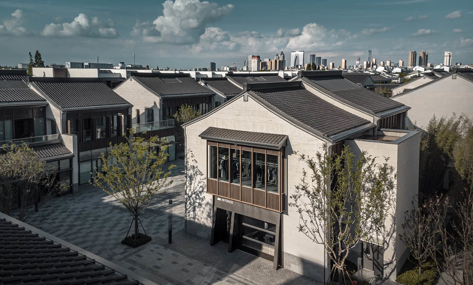 城市街道場景，現代建築具備混凝土立面與多扇窗戶，搭配兩旁種植樹木的鵝卵石街道