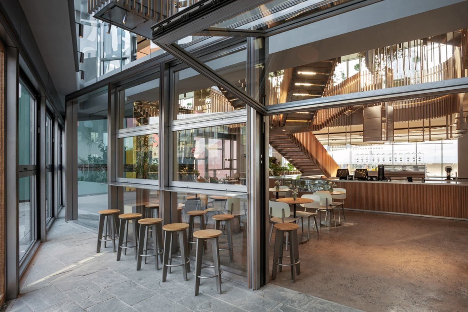Open restaurant interior with glass walls, wooden elements, and stainless steel accents, featuring dining tables, chairs, and potted plants