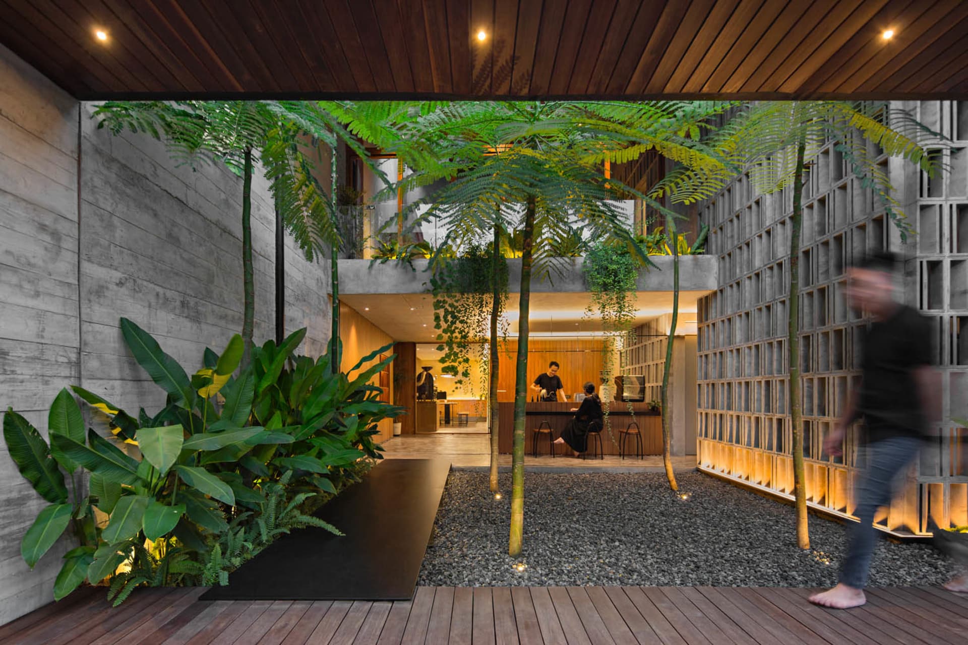 A modern cafe with a wooden floor, potted plants, and chairs arranged around dining tables, with people present in the scene