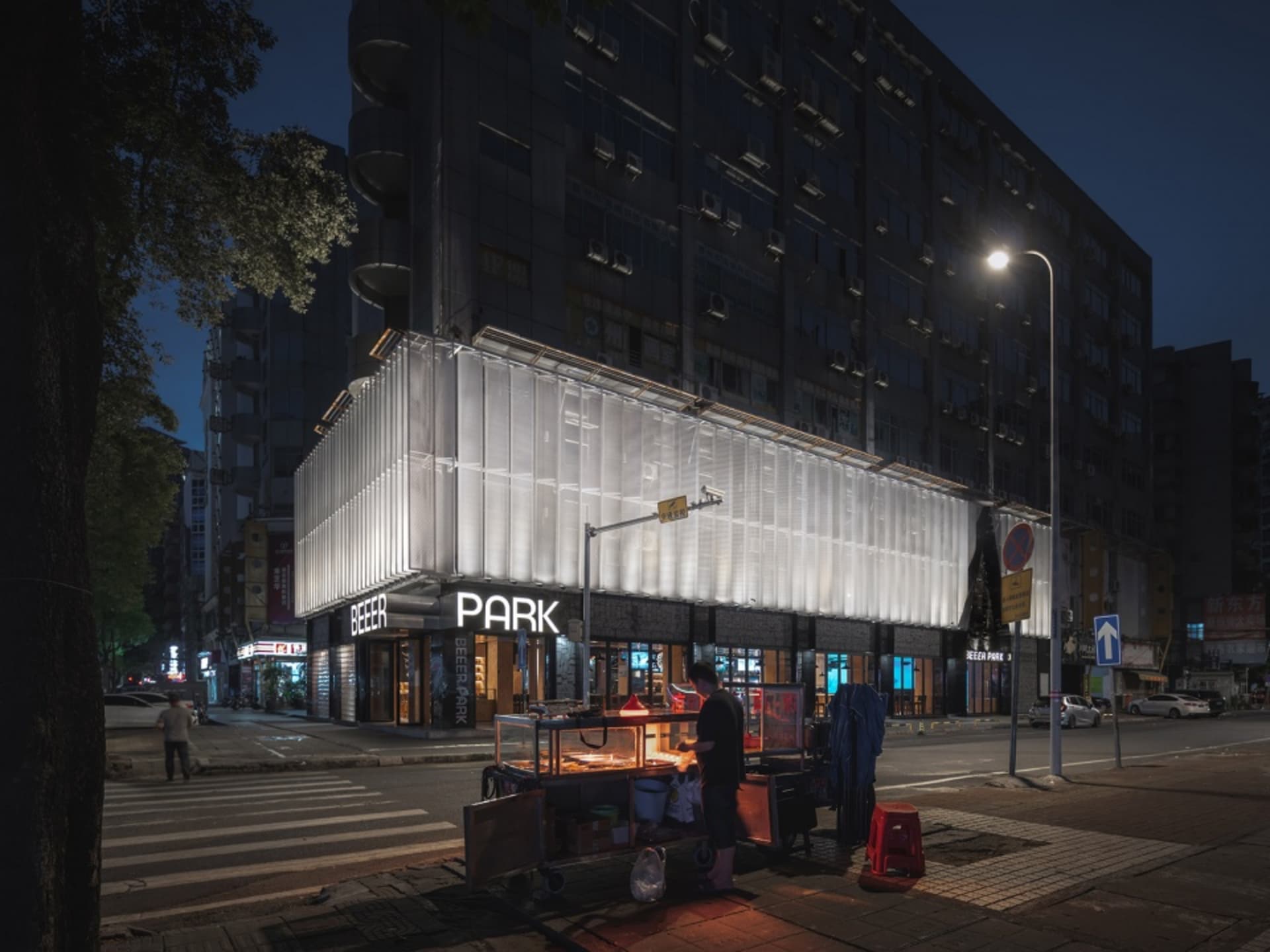 A bustling city street scene at night with a food cart and people walking by, with cars parked along the street and traffic lights in the distance