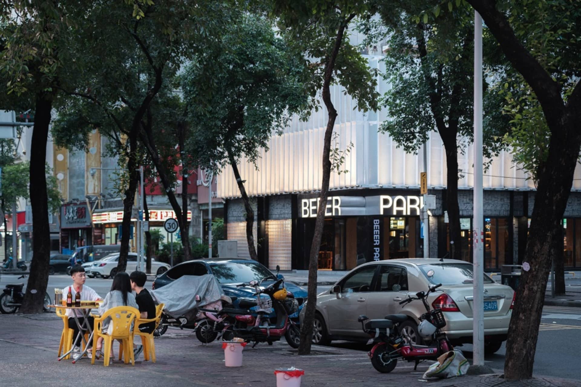 A city street scene with a white building, parked cars, motorcycles, and people walking around, with chairs placed at various locations
