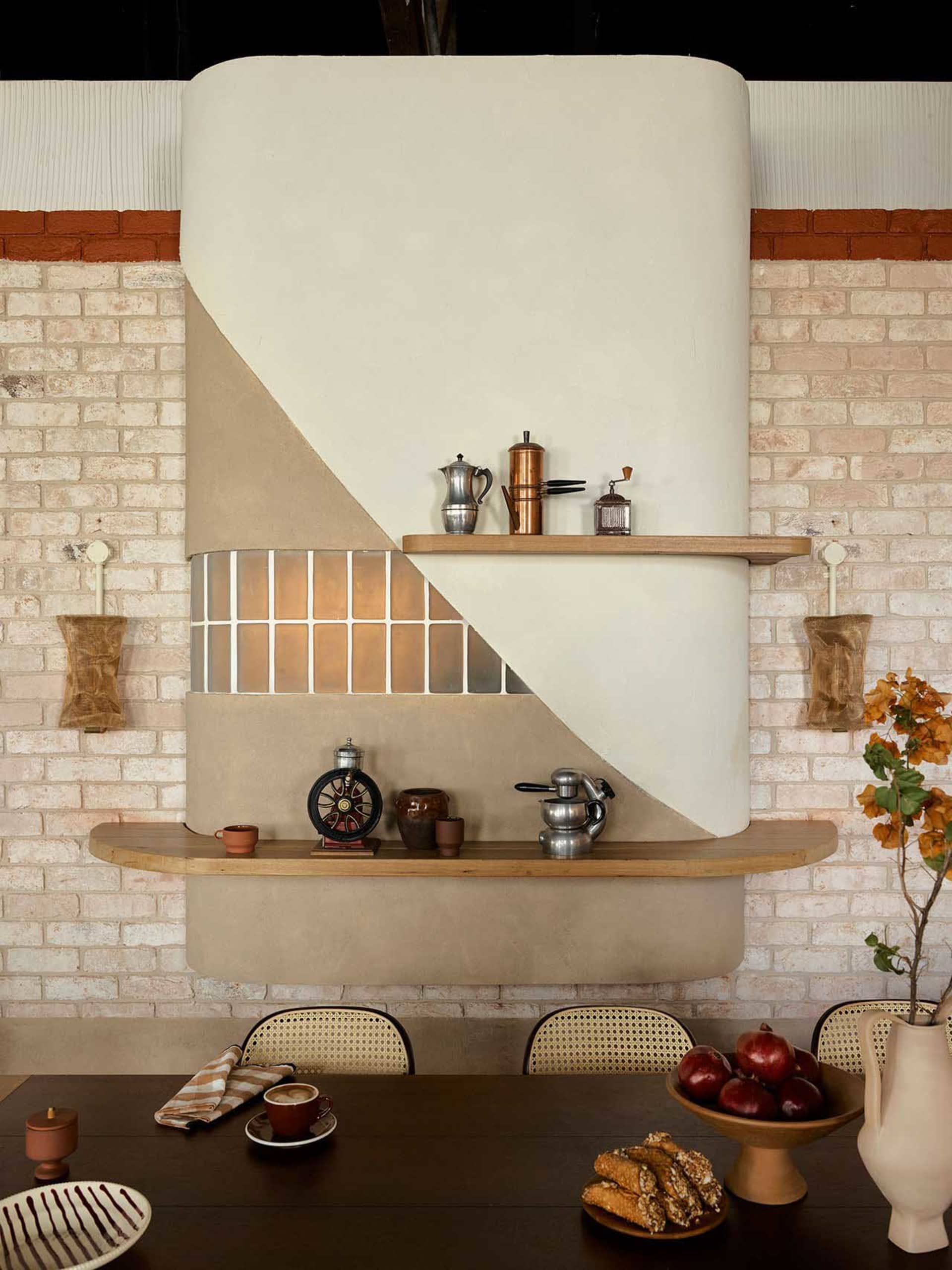 A warm and inviting kitchen space with a brick wall, white countertop, and various kitchen items scattered around, including chairs, cups, bowls, apples, and bottles, with a clock on the wall