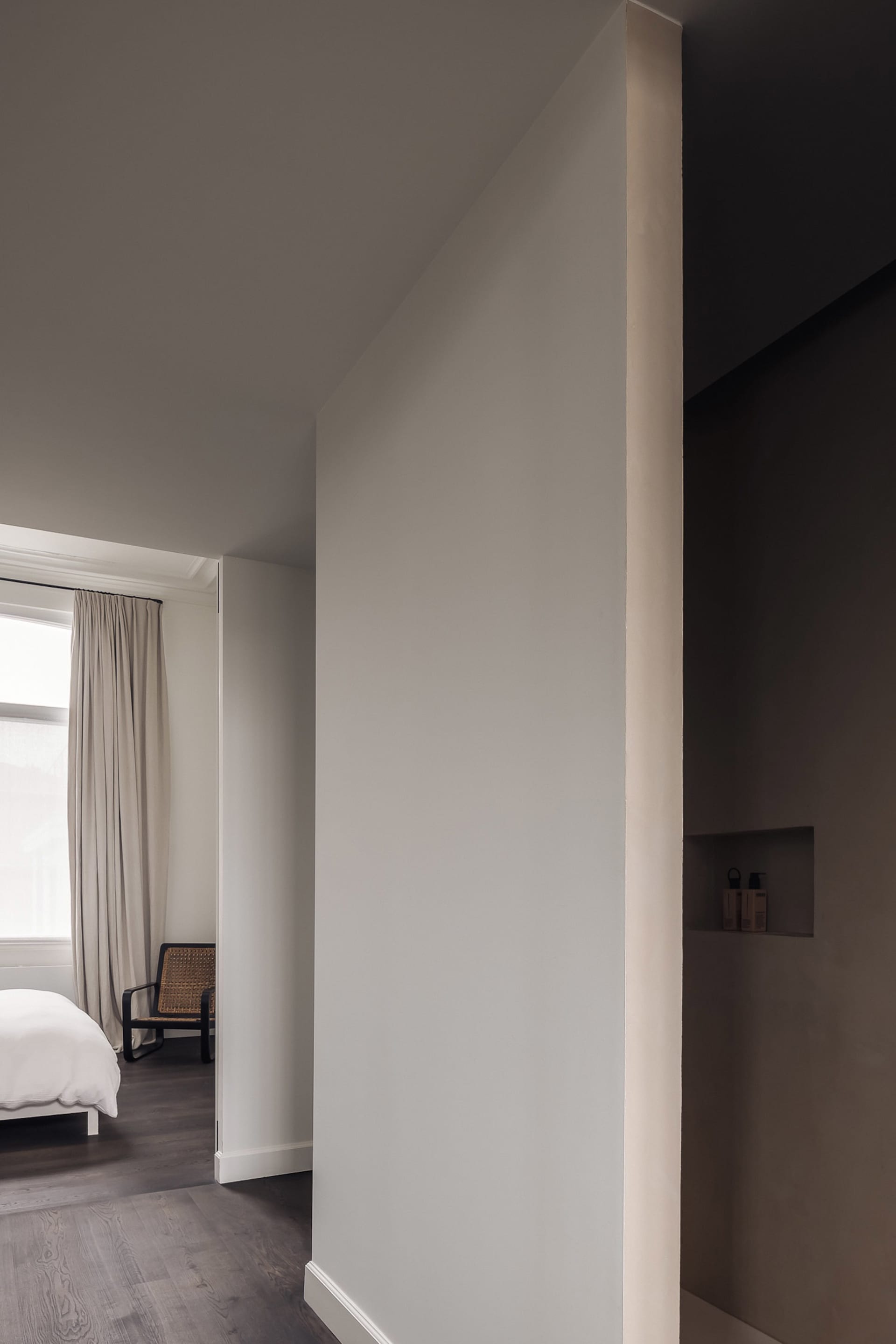 A dimly lit bedroom with a white bed and two chairs, surrounded by white walls and a minimalist aesthetic