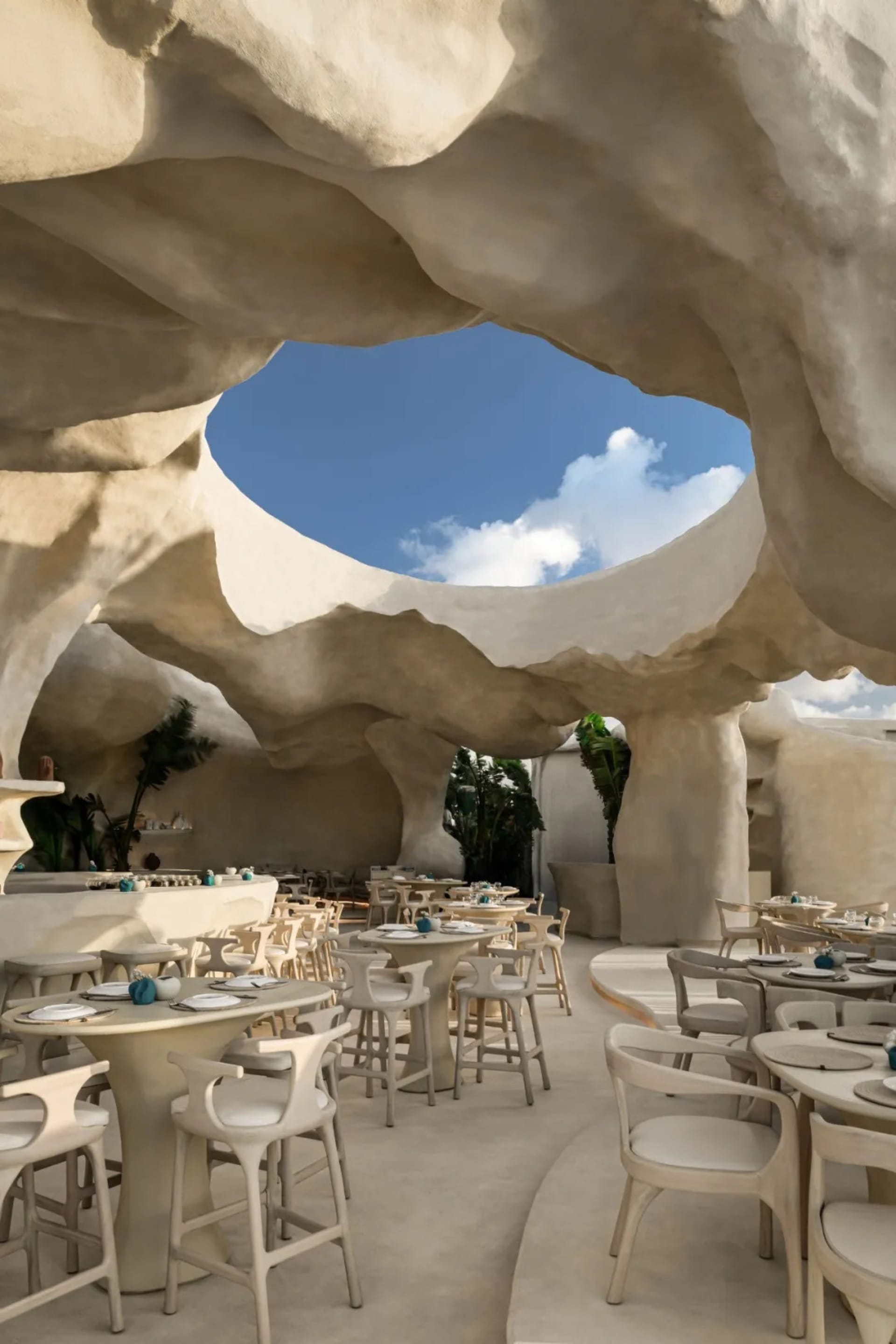 A spacious restaurant interior with white stone walls, multiple tables, chairs, and potted plants, creating a clean and inviting atmosphere