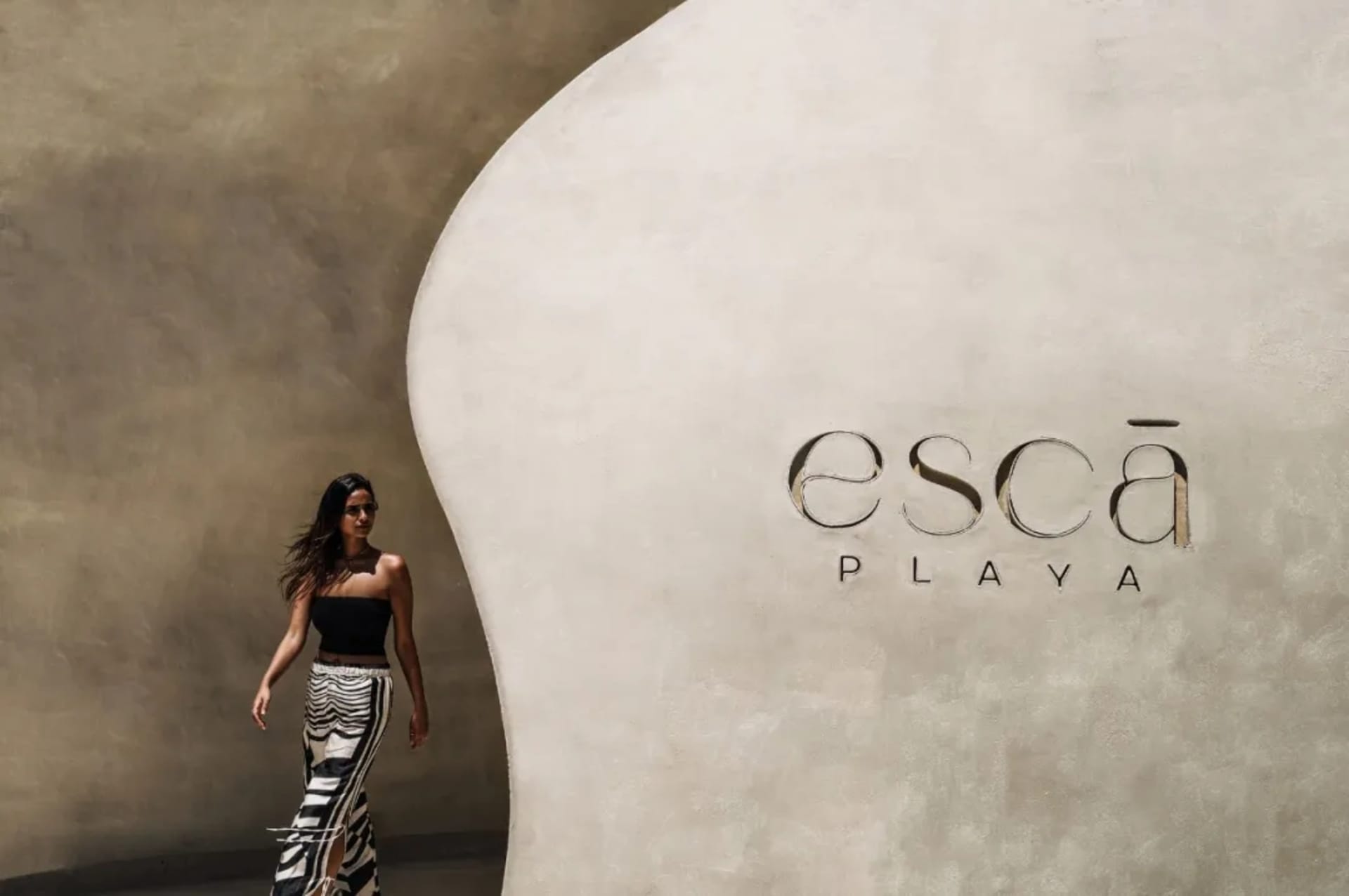 A woman in a black dress walks past a wall with an advertisement for Escapade Playa in a modern and stylish hotel lobby