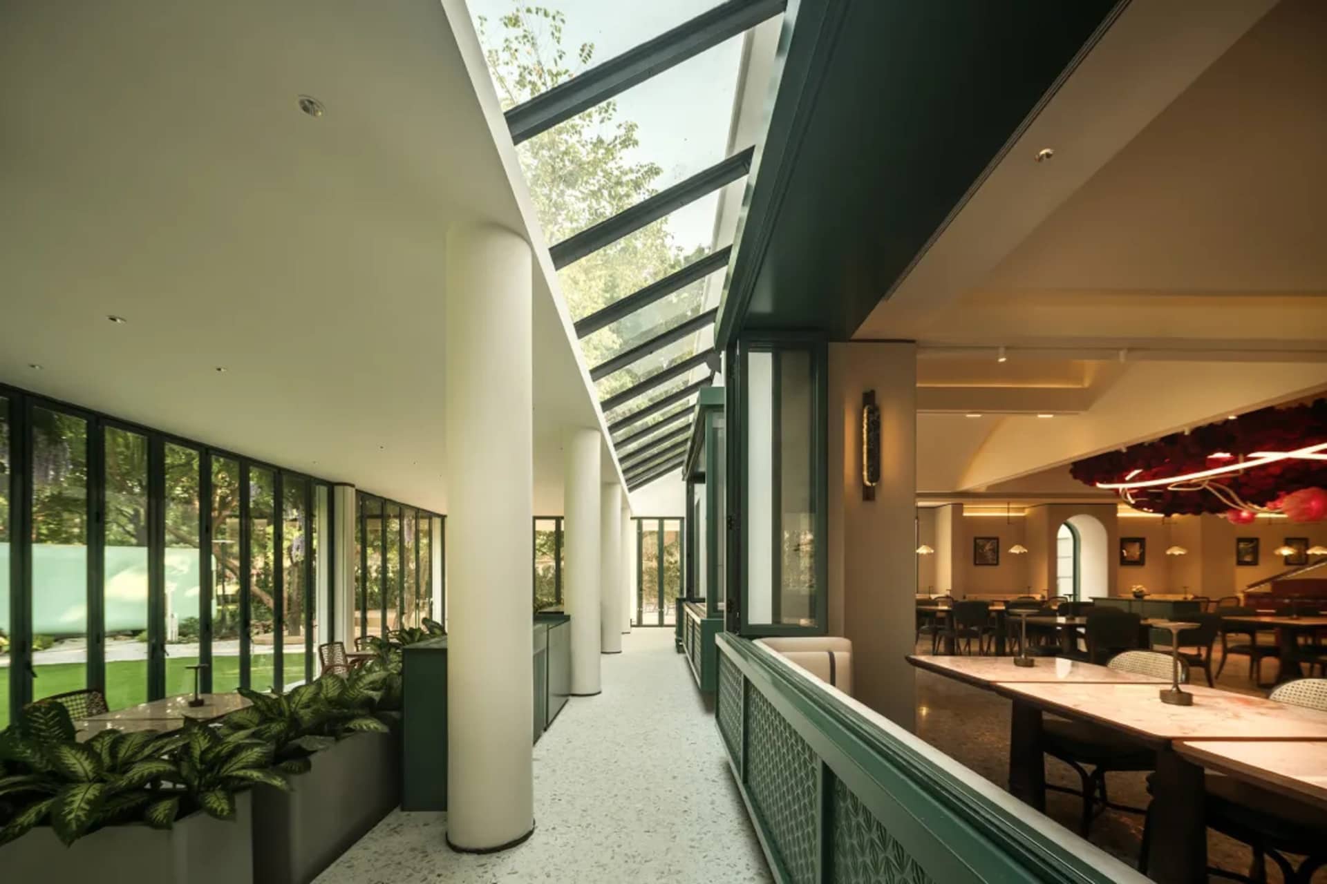 Modern restaurant interior with green walls, glass ceiling, and natural light, featuring dining tables and chairs, and potted plants