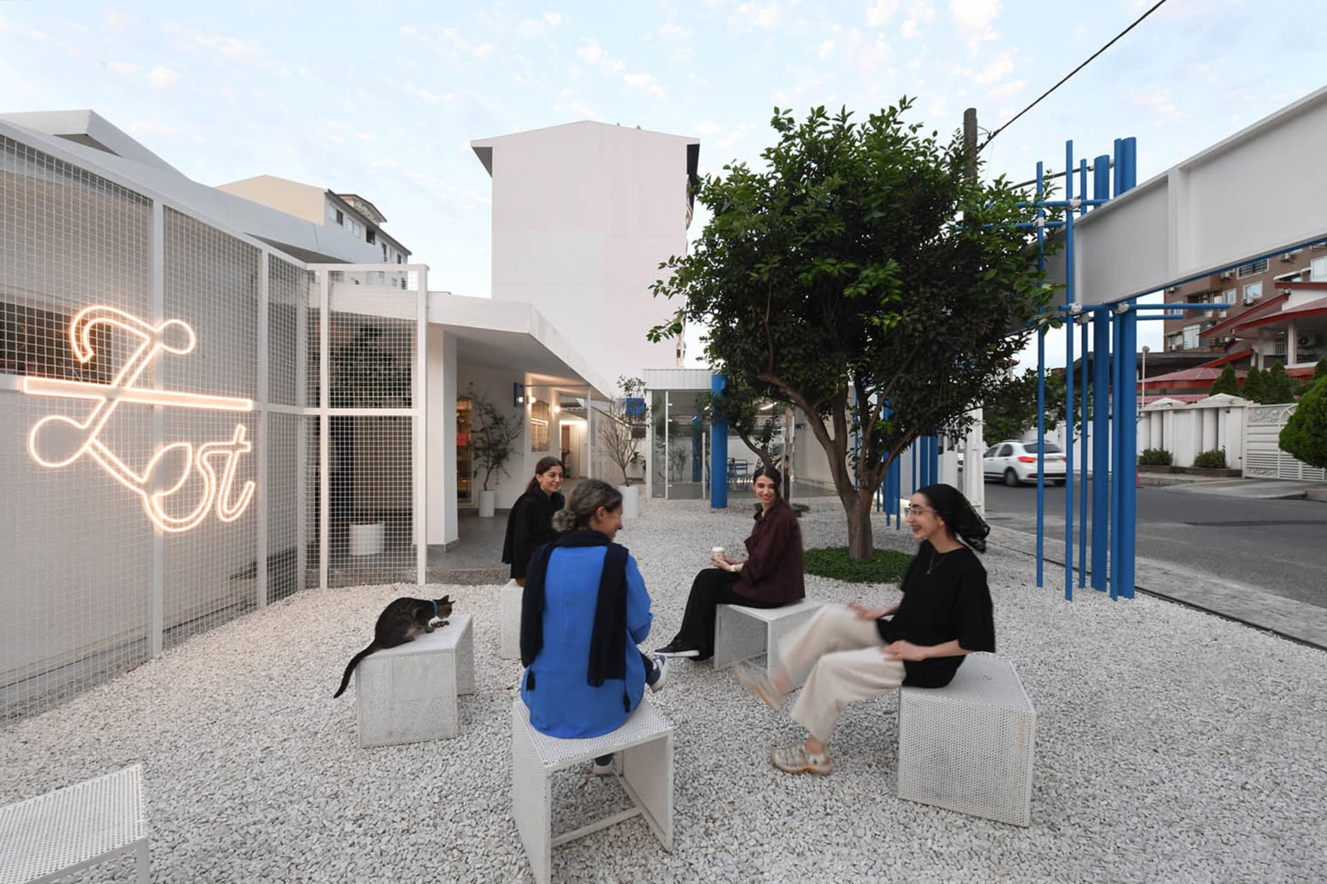 A casual outdoor gathering of people sitting on stools in front of a building with a white exterior, surrounded by a tree and enjoying each other's company