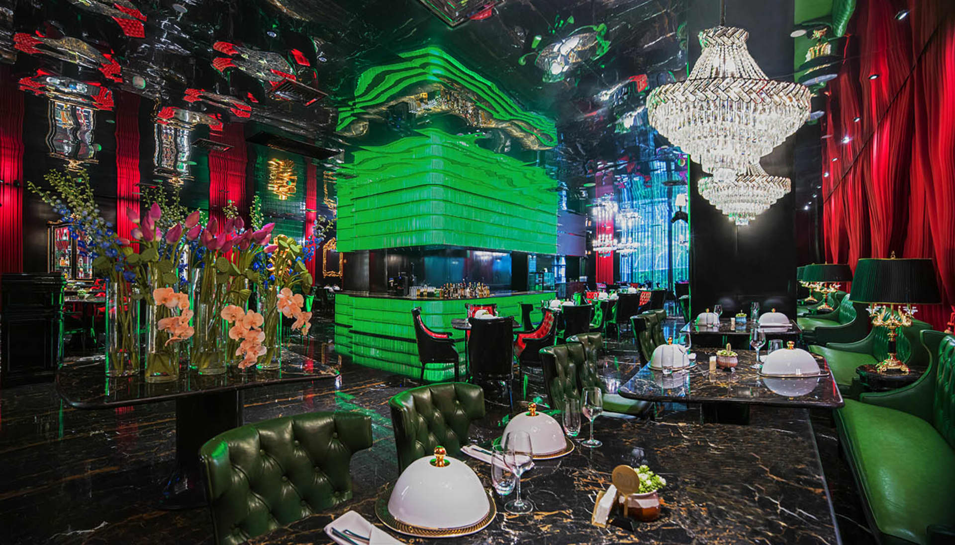 A modern restaurant with a large dining area featuring multiple tables, chairs, wine glasses, cups, and bowls, set against a backdrop of green walls