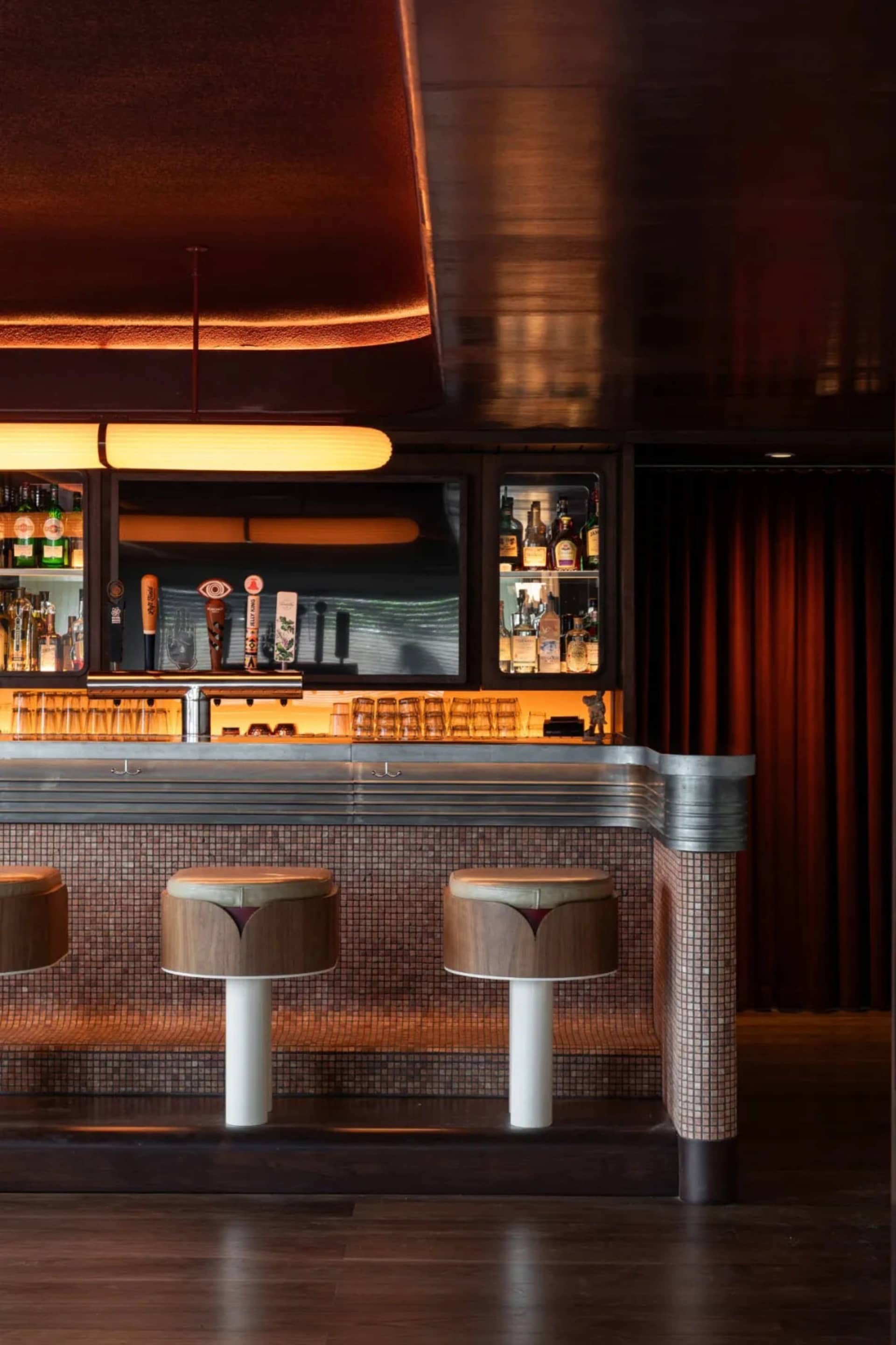 A cozy bar area with a wooden counter, stools, and a TV, surrounded by bottles and creating a warm atmosphere