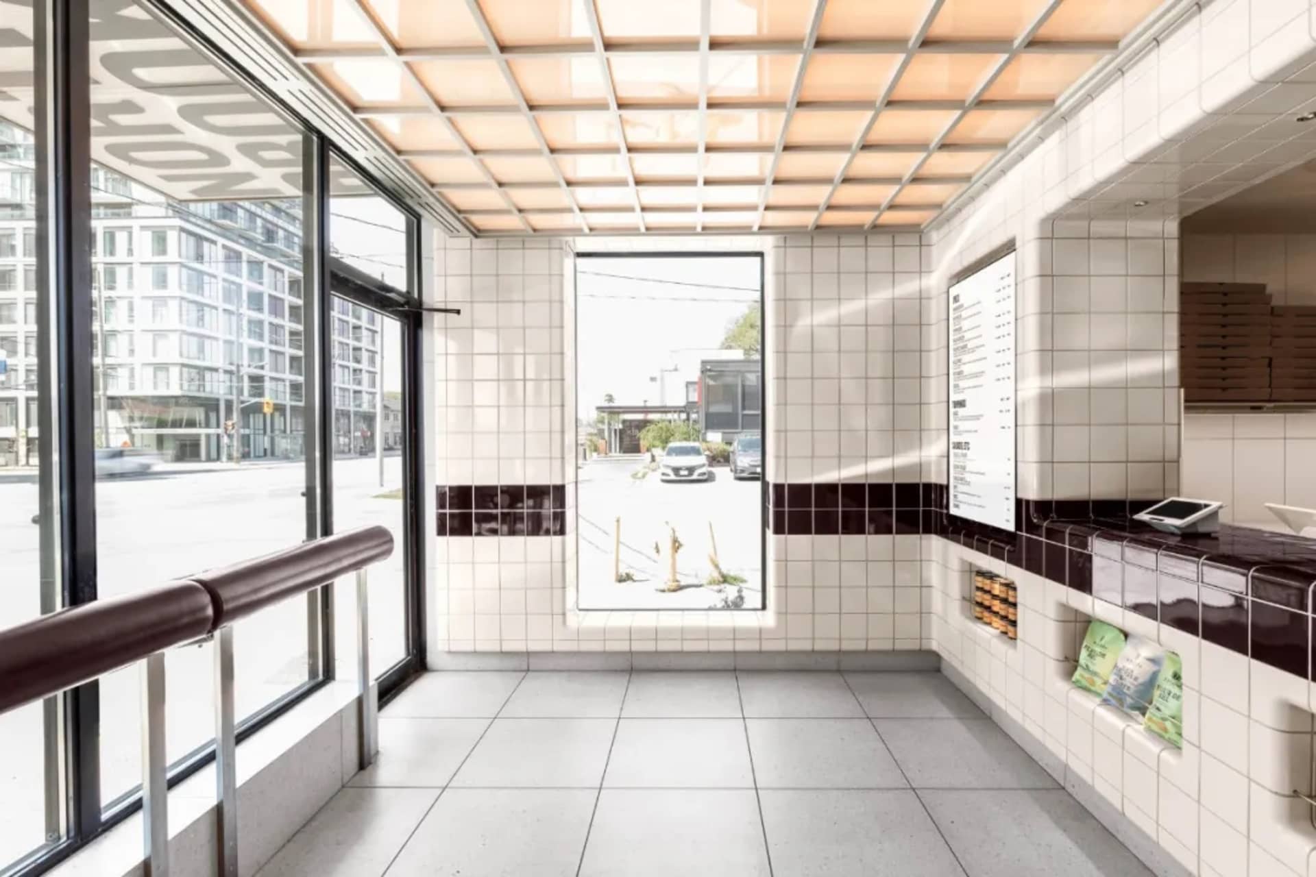 A modern interior space with a tiled floor, walls, and a large window allowing sunlight to enter, featuring two benches and a view of a car outside