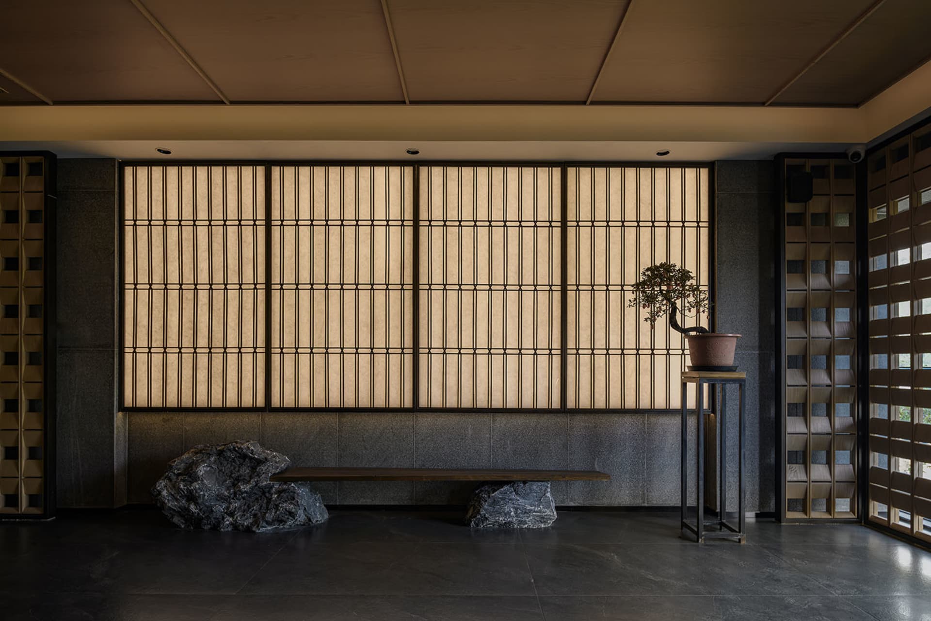 Asian-inspired living area with a large window, wooden blinds, potted plants, a bench, and a vase, creating a calm and serene atmosphere