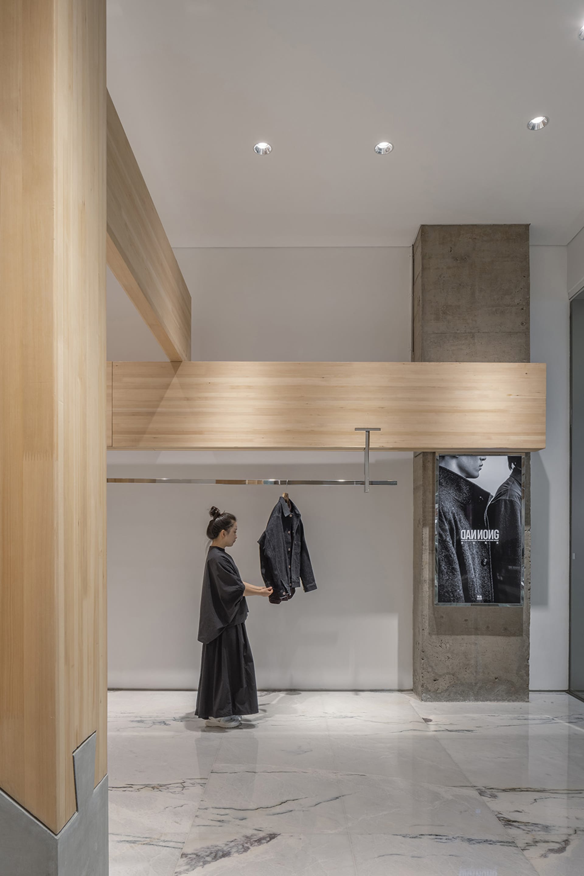 A woman holds up a black jacket in a well-stocked retail store with a concrete wall and various clothing items on display