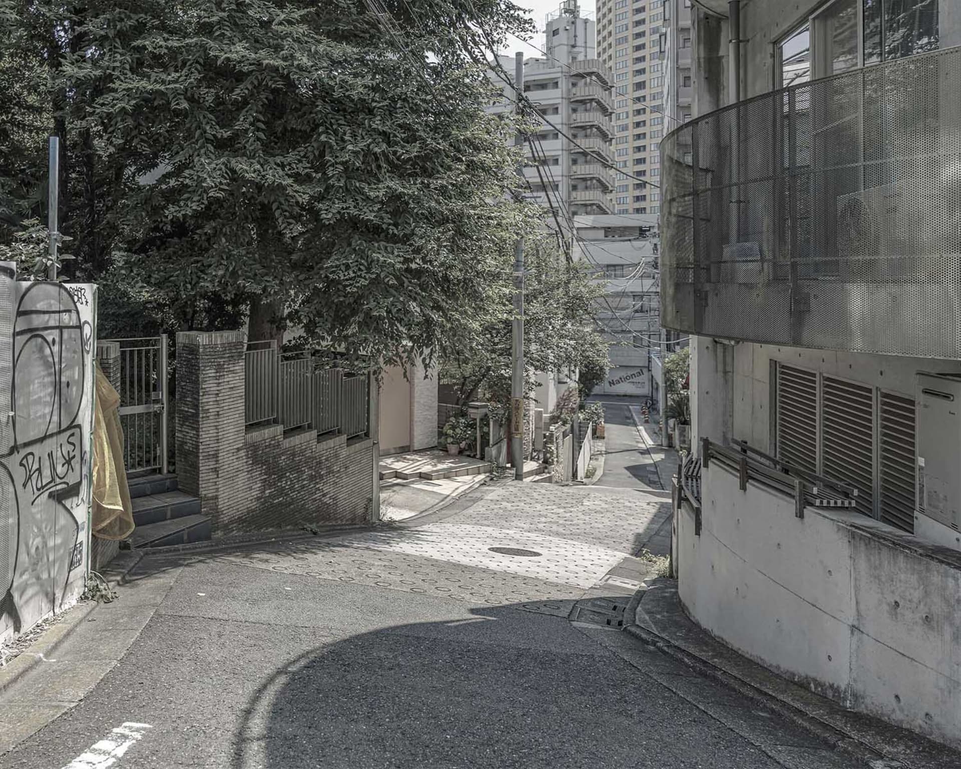 A calm urban street scene with a tall building and a tree on the opposite side, two people walking in the distance