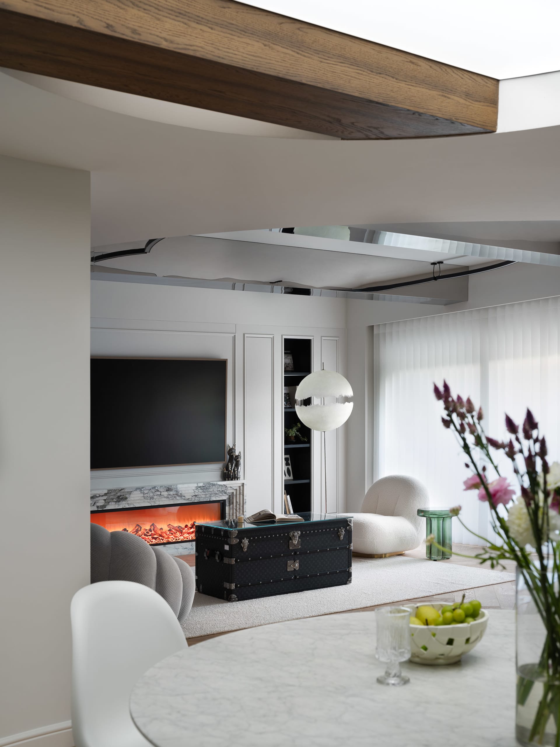 Cozy living space with a fireplace, a couch, chairs, and a dining table, featuring a mix of white and brown tones, with vases and apples adding a touch of freshness