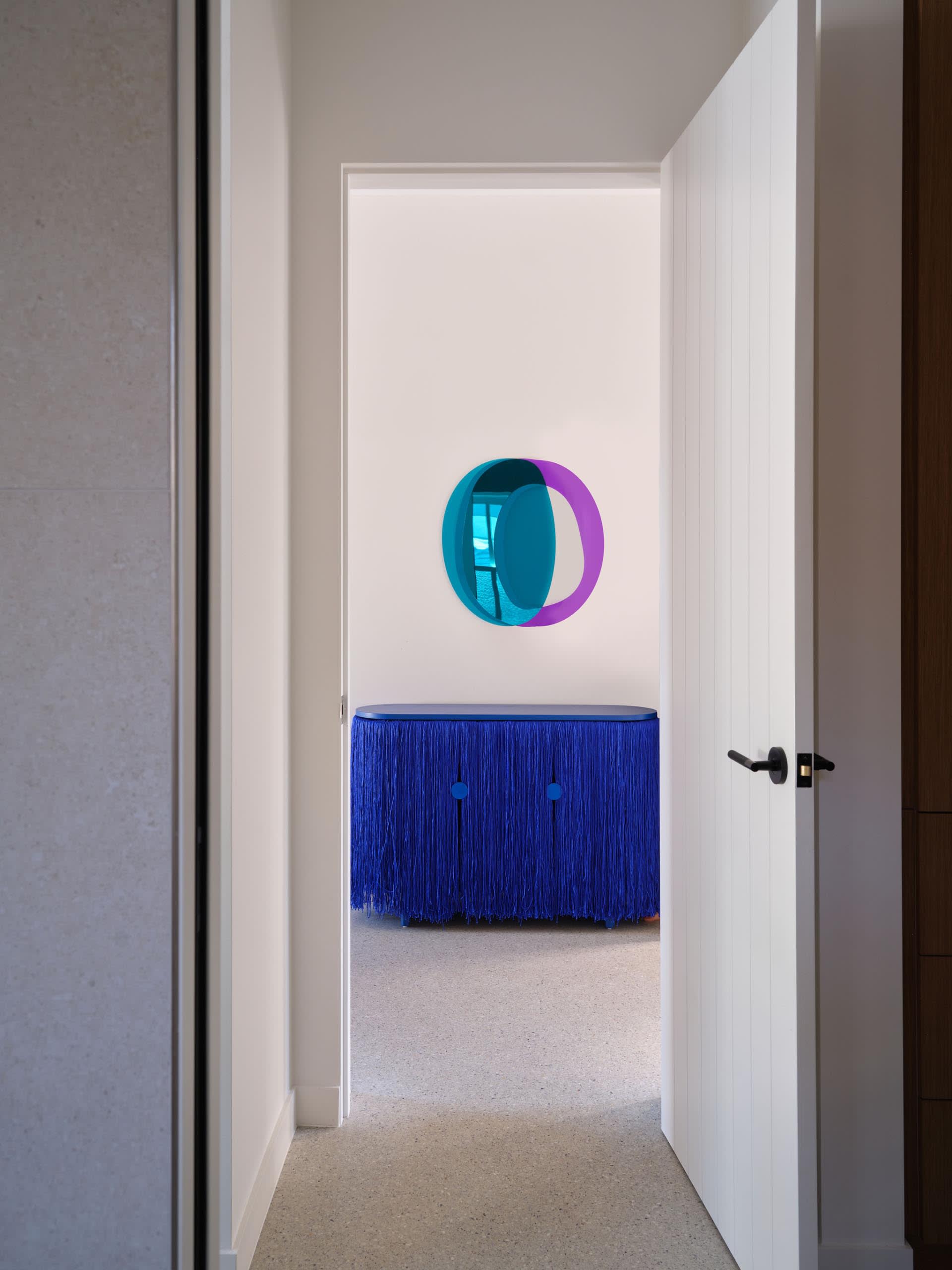 Cozy living room with a blue couch in front of a mirror with a purple circle design, two chairs, and a doorway leading to another room