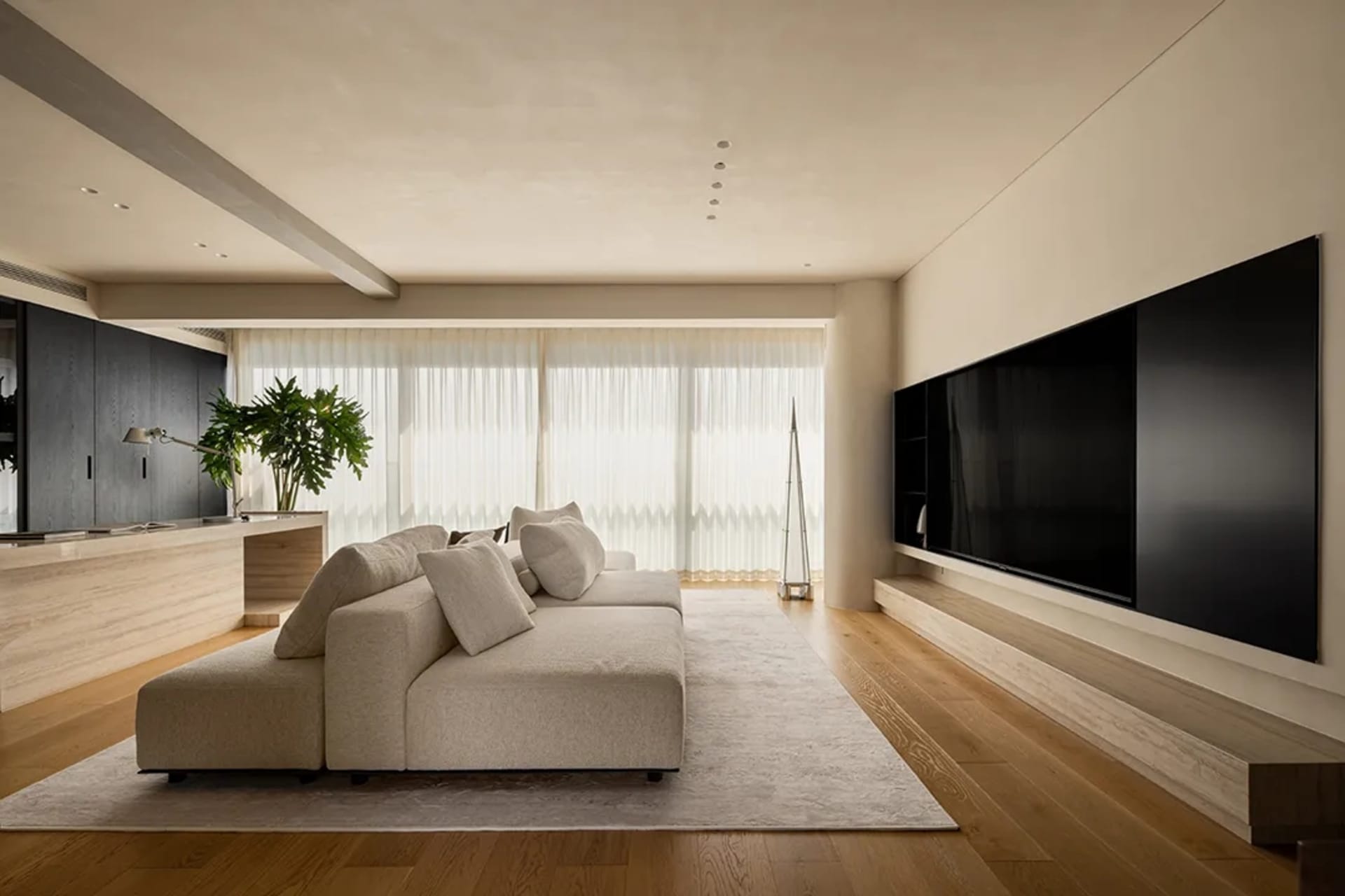 A spacious living room with a large couch, two chairs, a mounted television, and a potted plant, set against a backdrop of white walls and natural light