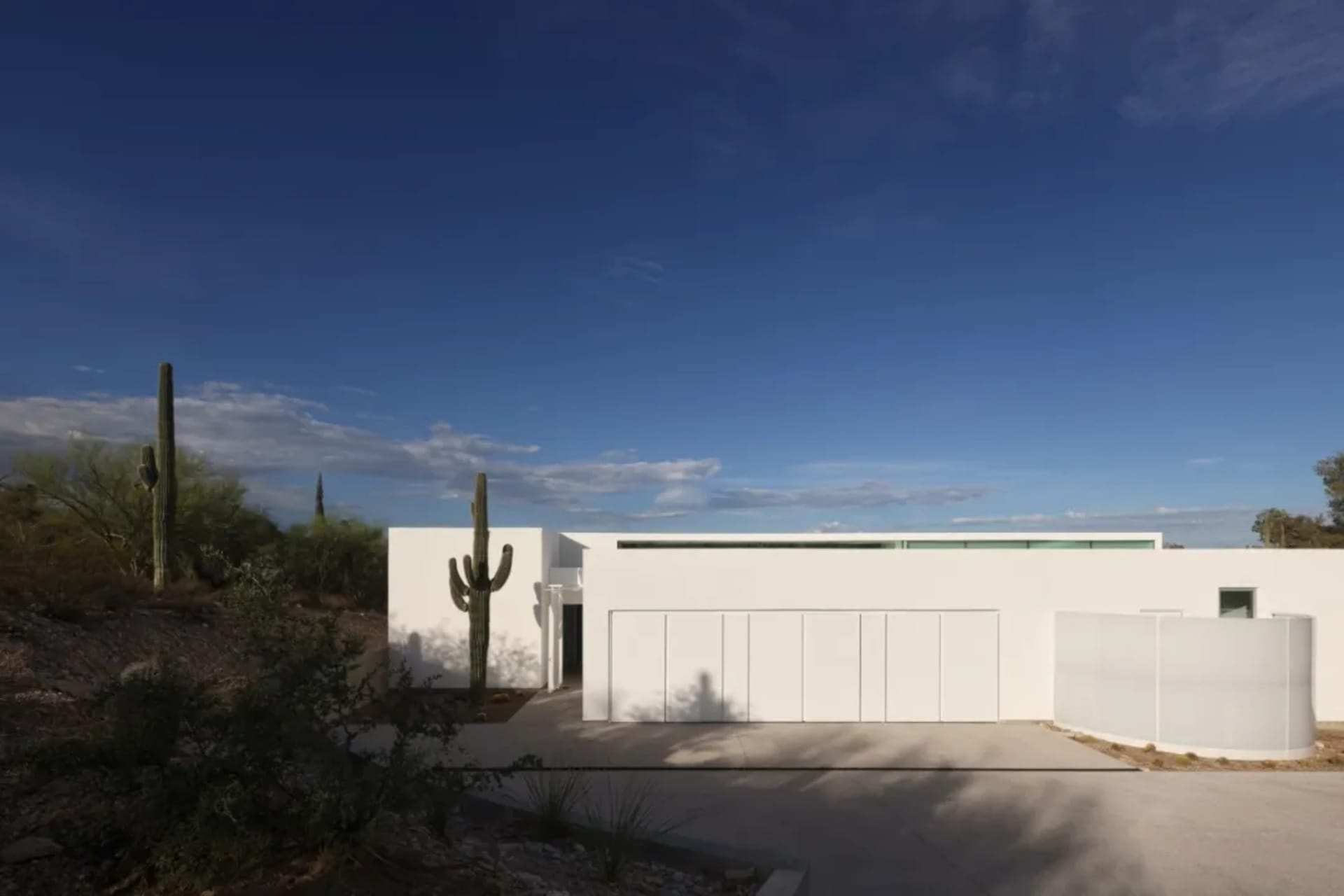 Exterior view of a modern white house with a large garage, two cacti, and a large window, bathed in sunlight