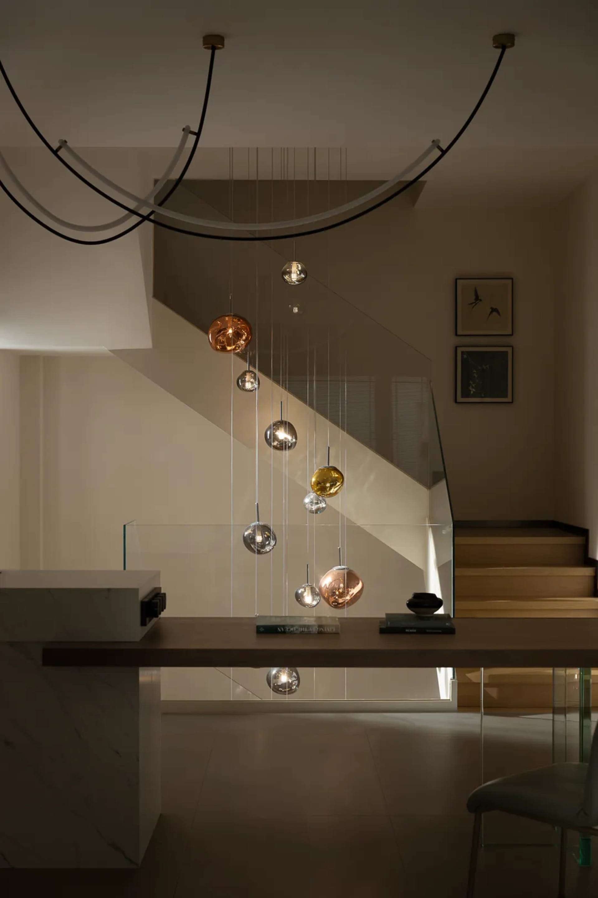 Elegant staircase with glass railing, white wall, and dining table with chairs and books, creating a warm and sophisticated atmosphere