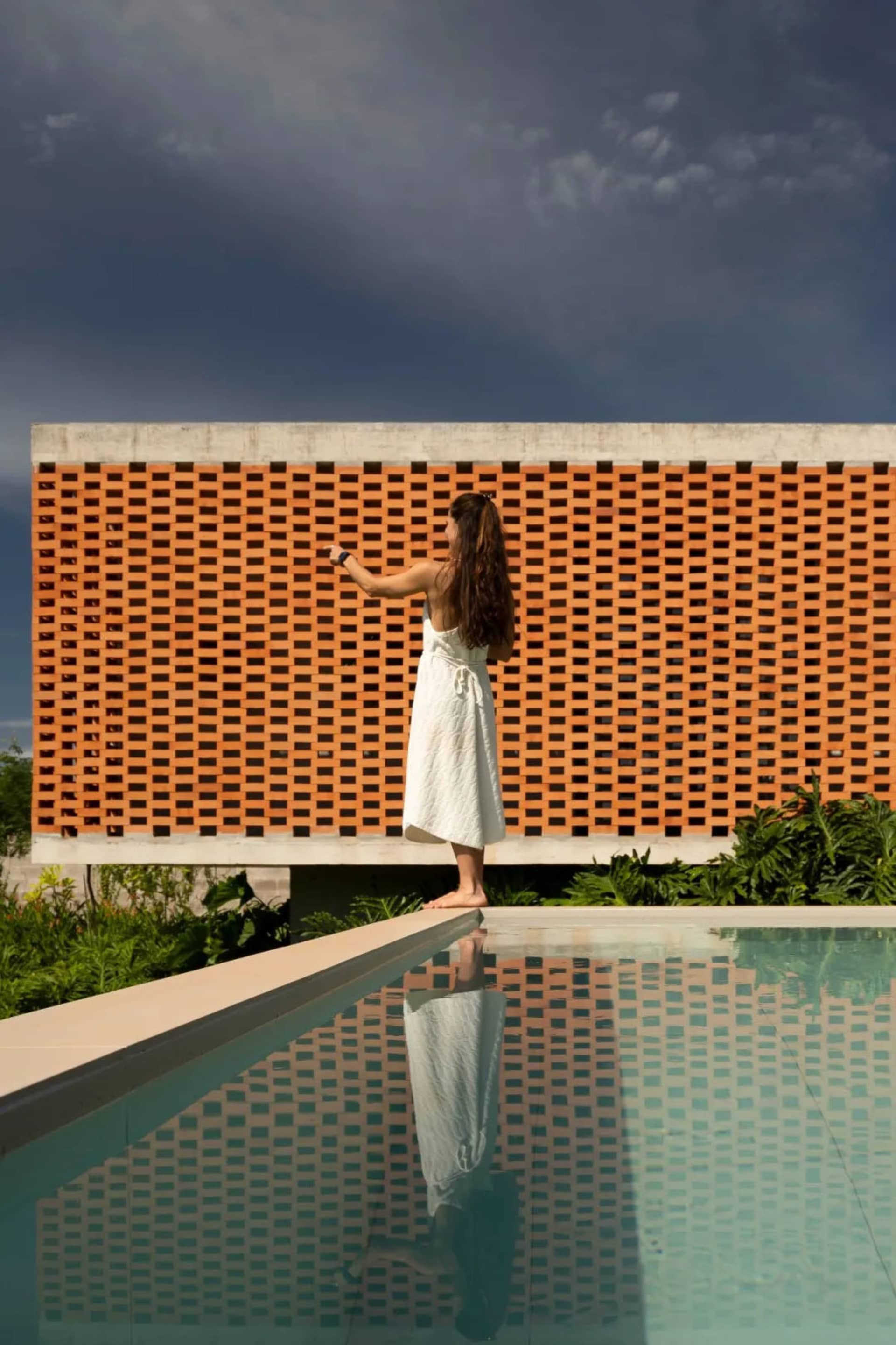 A woman stands near a pool, taking a photo of the surrounding area with a reflective pool surface adding depth to the scene