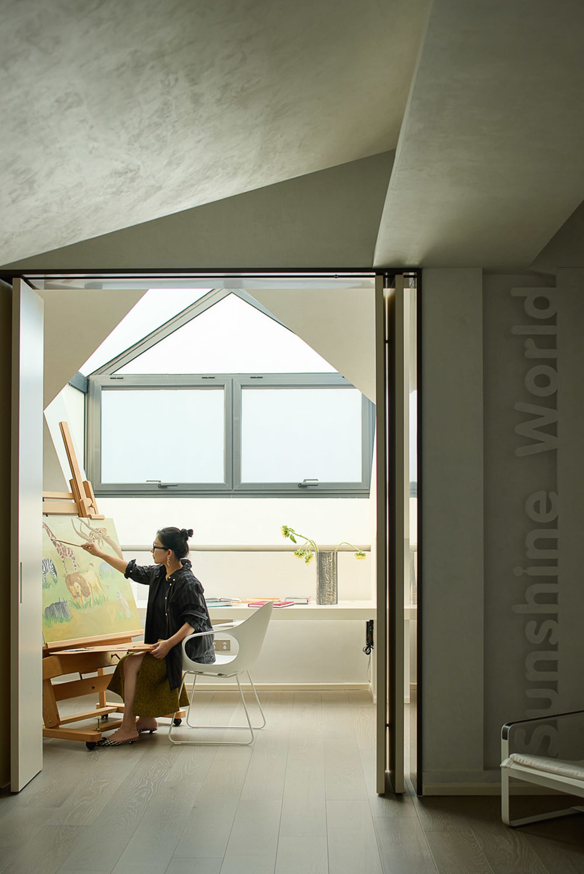 An artist works in a well-lit studio with a desk, chairs, books, and a potted plant, surrounded by natural light from windows
