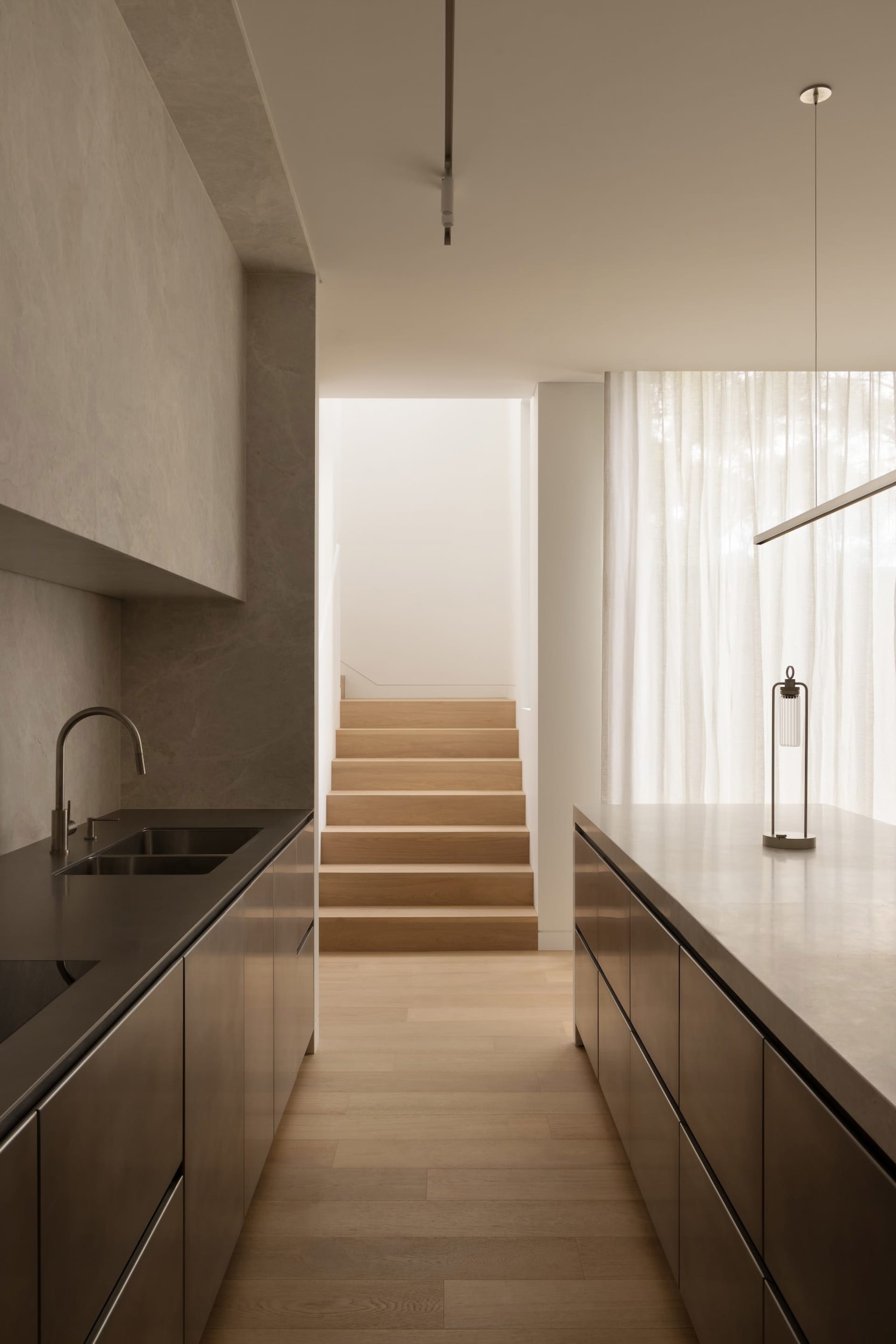 A modern kitchen with wooden floors and cabinets, featuring two sinks and a staircase leading up to another level, with a bright and airy ambiance