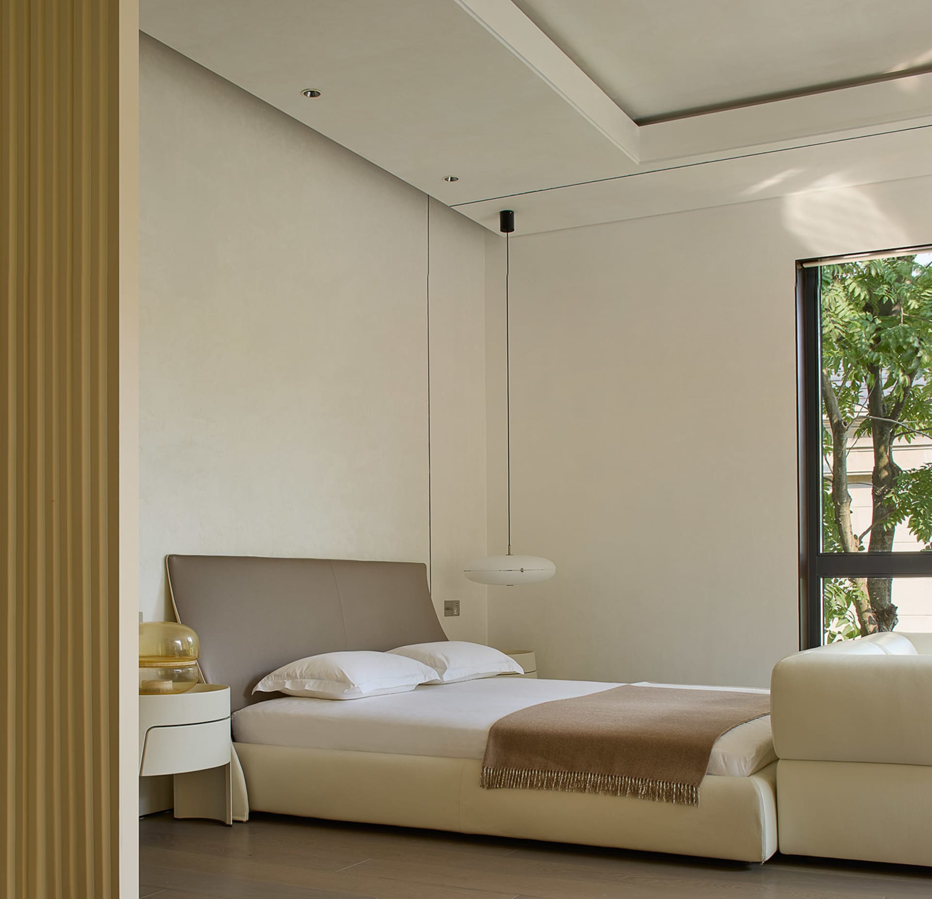 A large bedroom with a minimalist design, featuring a white couch next to a window, two beds, and a chair, giving a calm and inviting atmosphere
