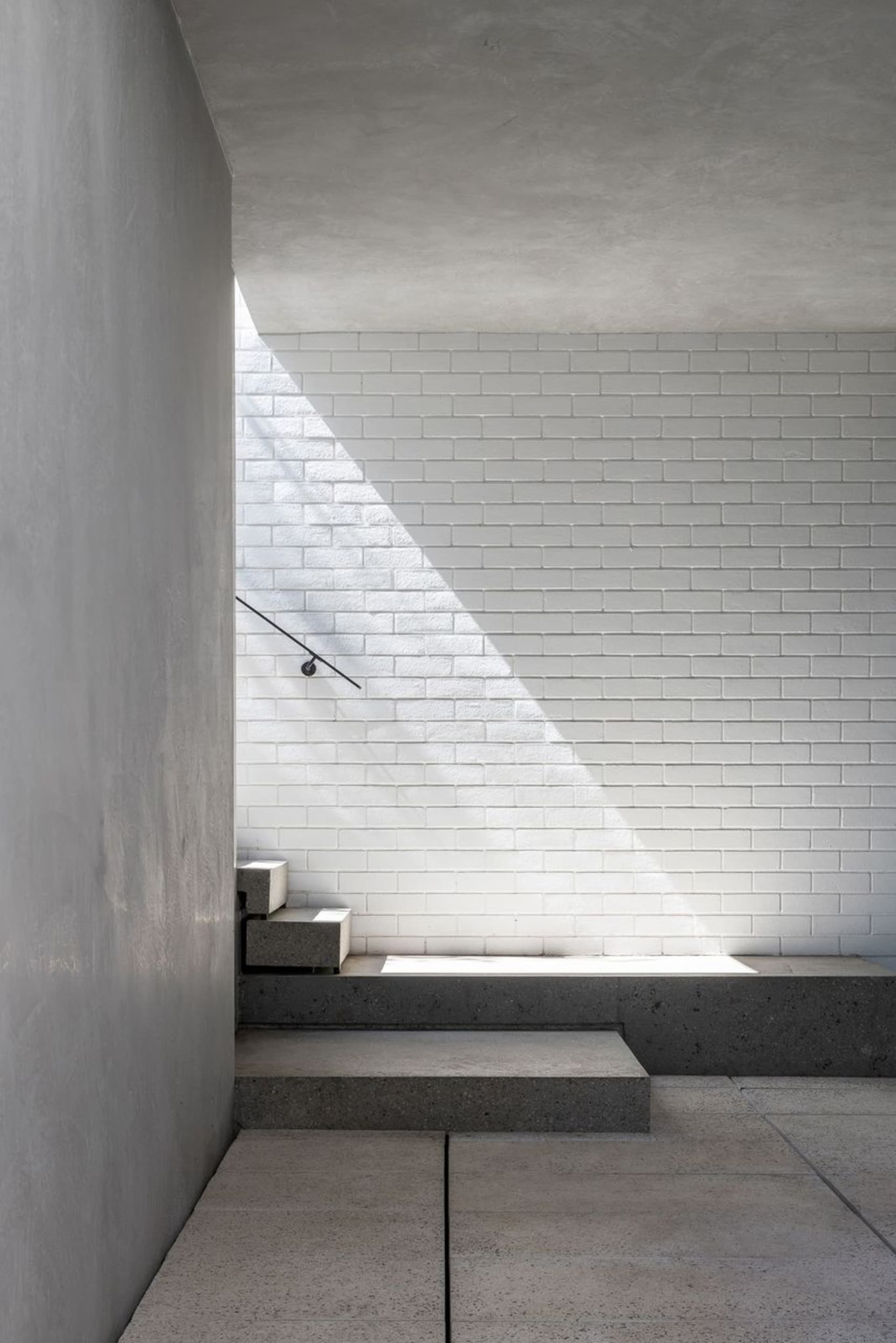 A minimalist staircase with a handrail leads to a white brick wall, set against a possibly residential or commercial building exterior