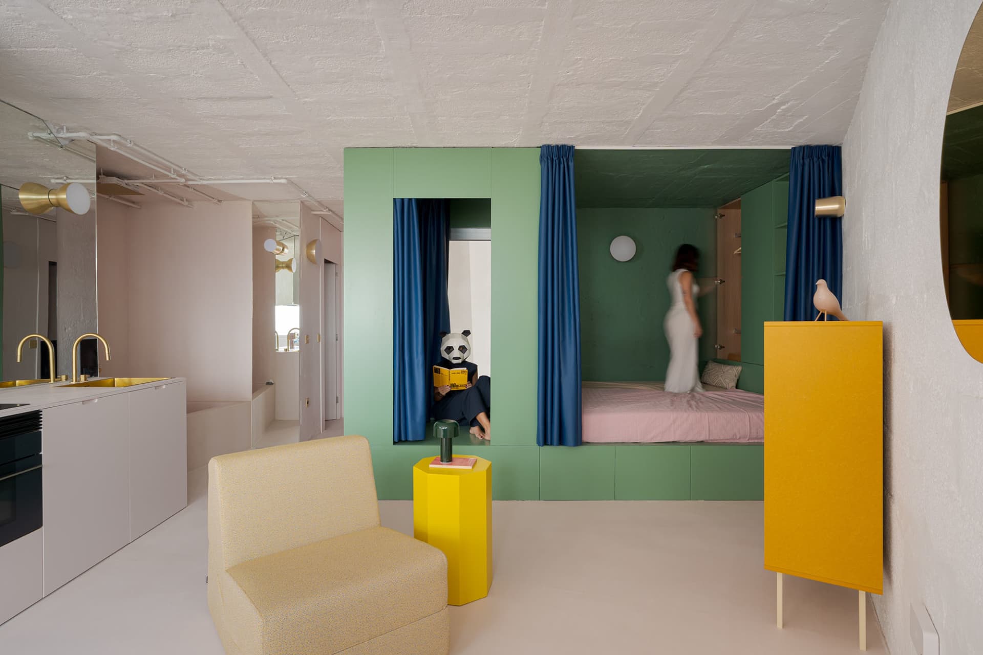 Cozy bedroom with green walls, yellow accents, two beds, a chair, a couch, and a person standing in the middle of the room