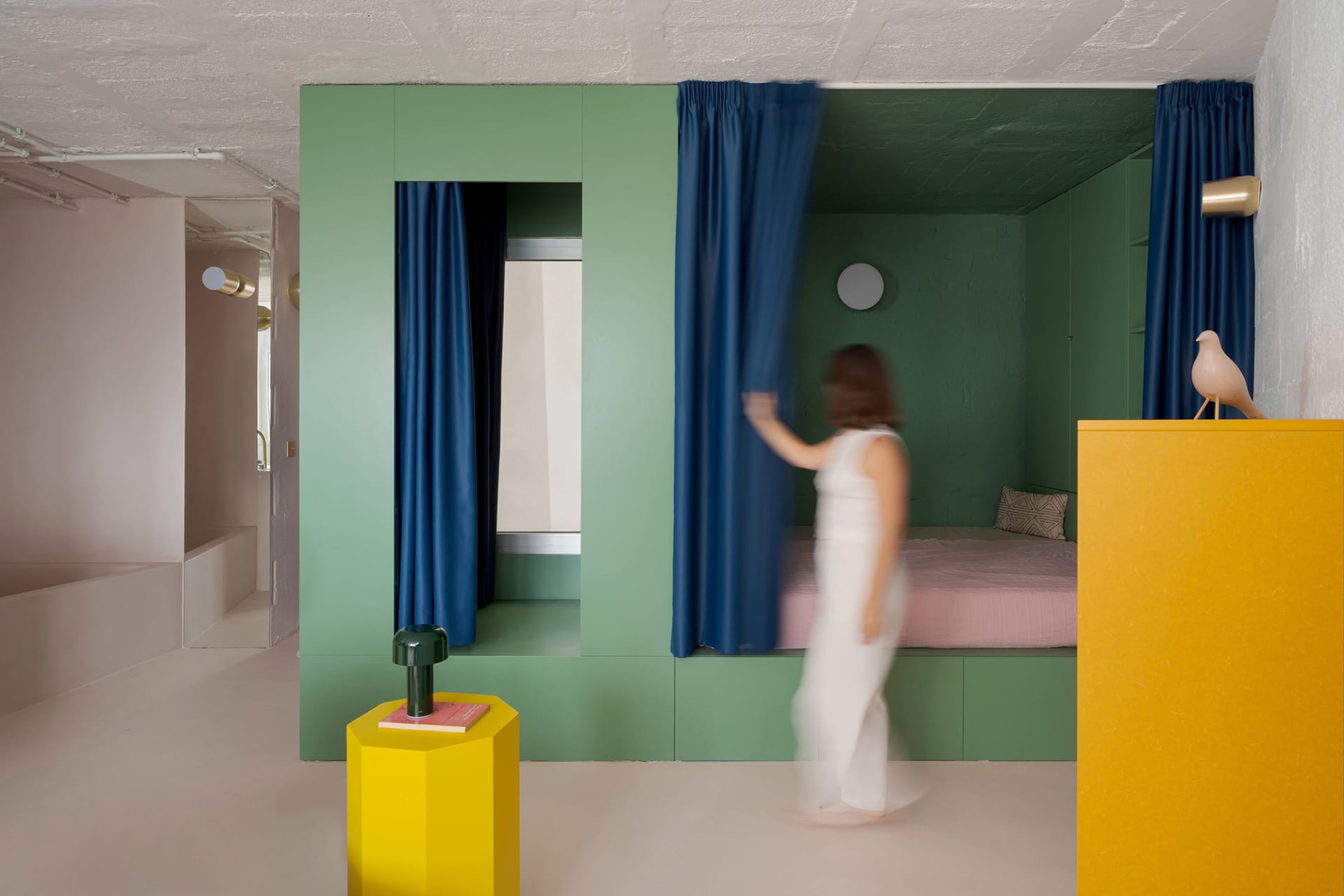 A woman walks through a calm and inviting bedroom in a green house with a bed, chairs, and a book, surrounded by white furniture and a serene atmosphere