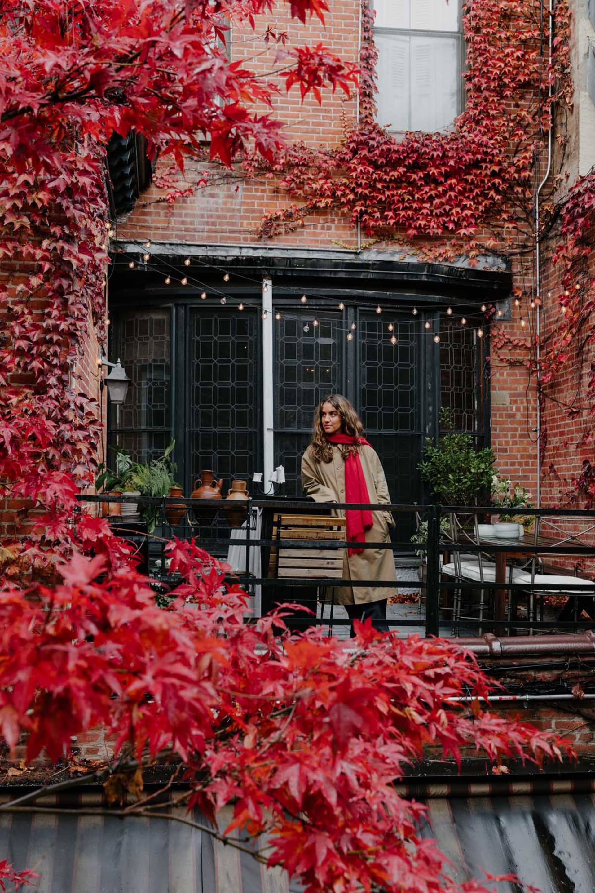 A woman poses on a balcony with a red brick wall and potted plants in the background