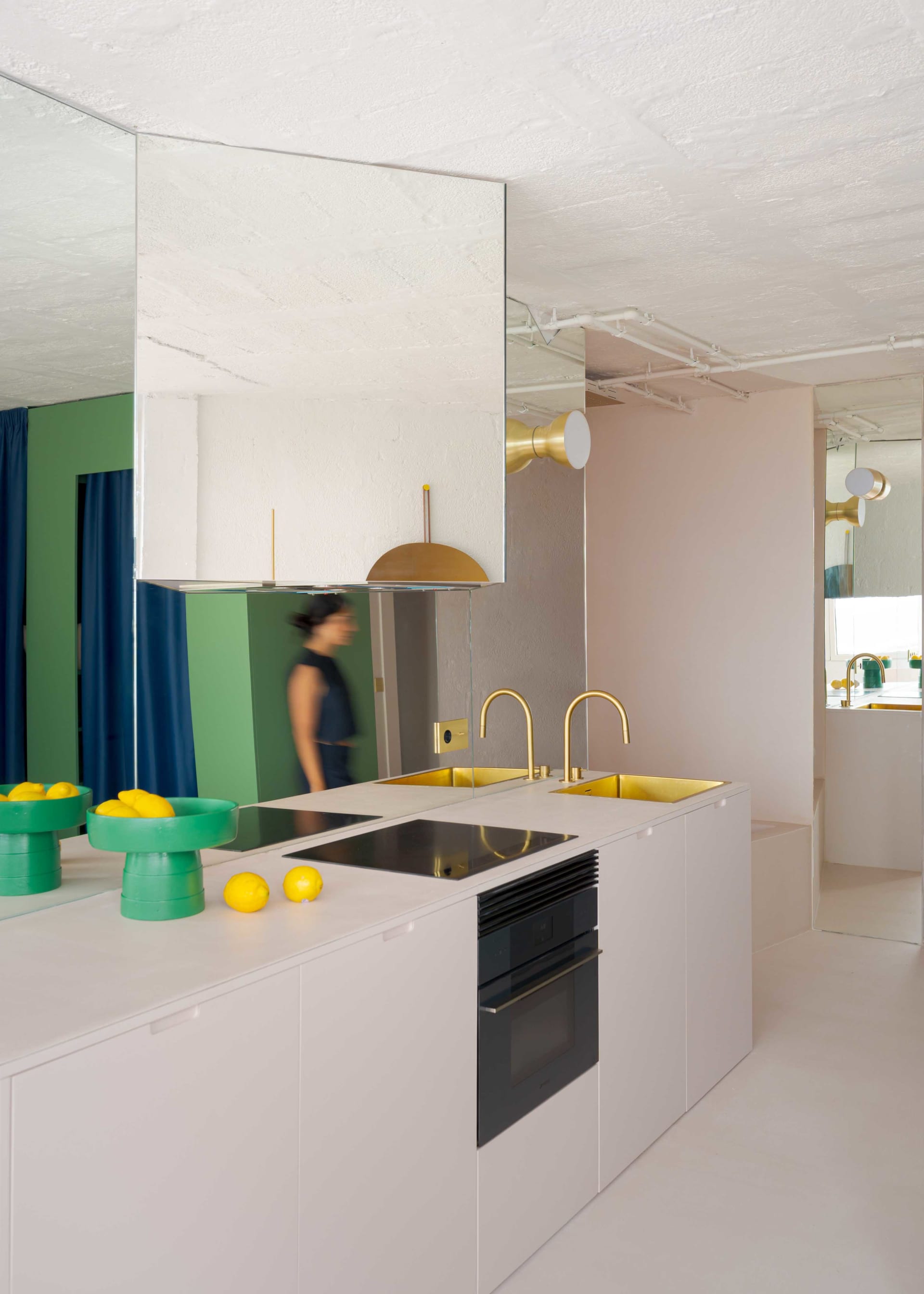 A kitchen with white cabinets, two sinks, and various fruits and bowls, with a woman walking through the space