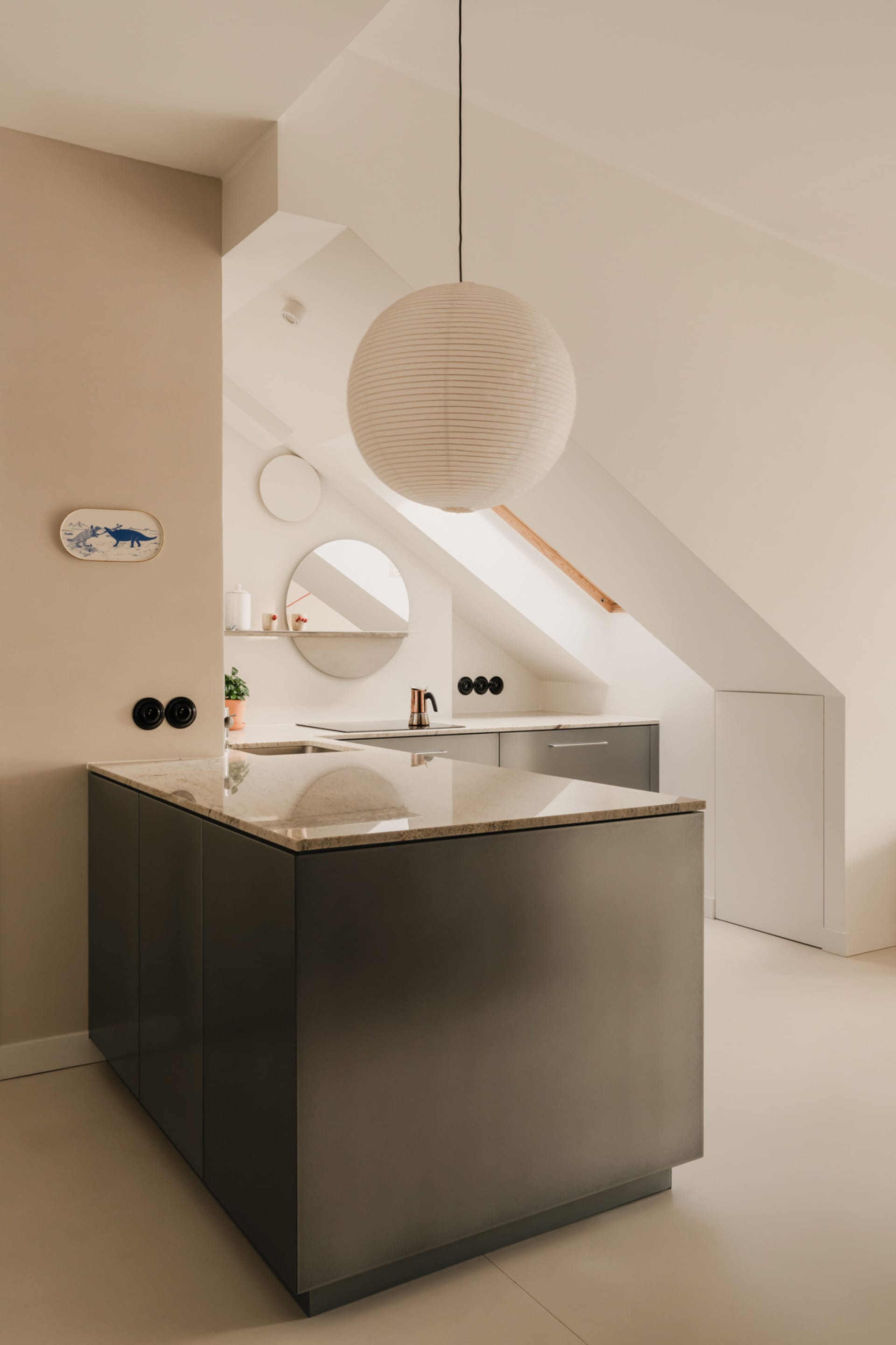 Modern kitchen with white walls and floors, featuring a large island, countertops, cabinets, sinks, a hanging lamp, and potted plants