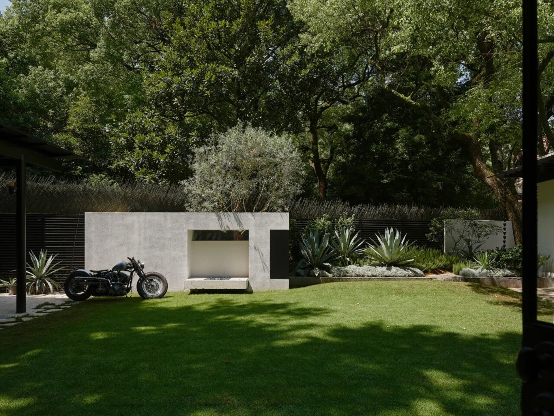 An outdoor living area with a motorcycle, potted plants, and benches, surrounded by greenery and concrete elements