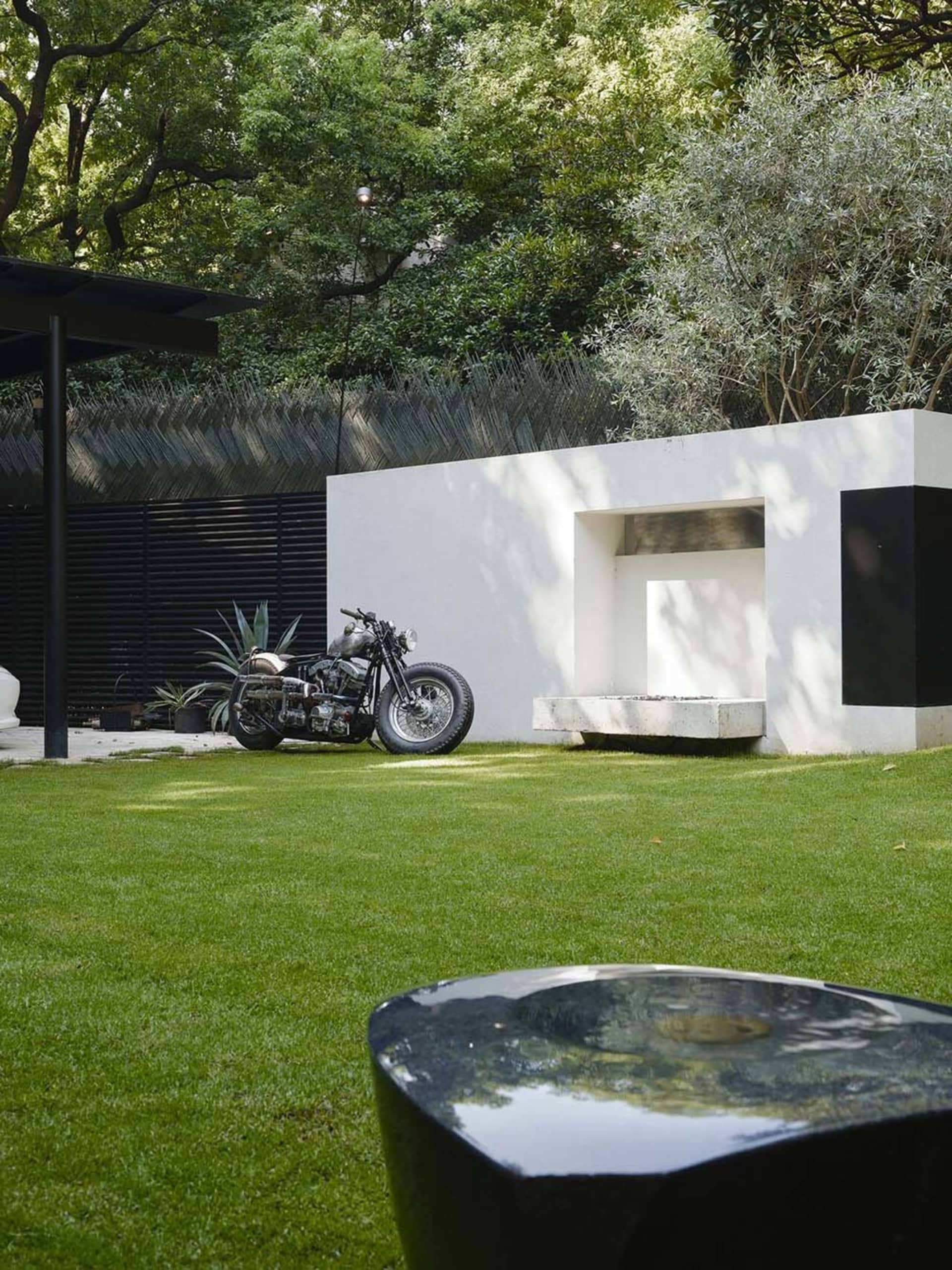 A relaxed outdoor area with a motorcycle parked on a grassy lawn in front of a white building, featuring two benches and a pleasant atmosphere