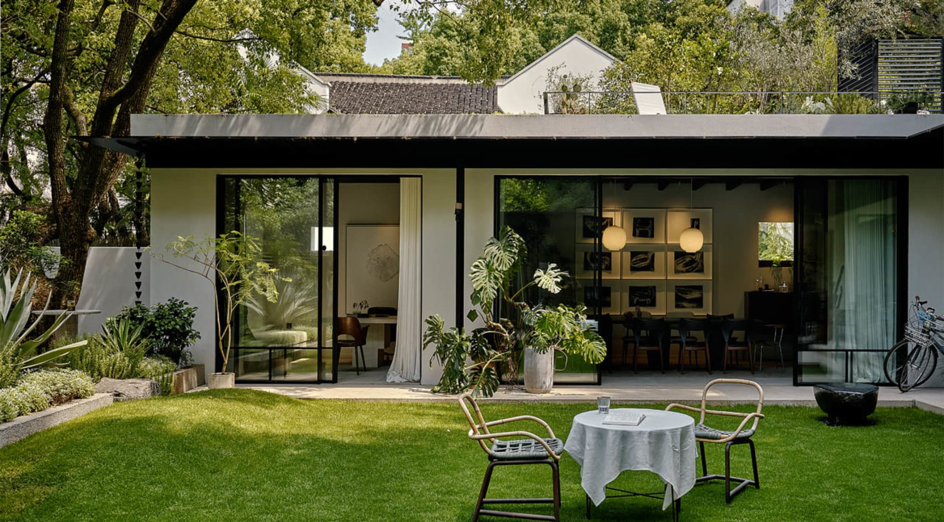 Open modern living space with white walls, green grass, and natural furniture, featuring a dining table and multiple chairs, a potted plant, and a contemporary atmosphere