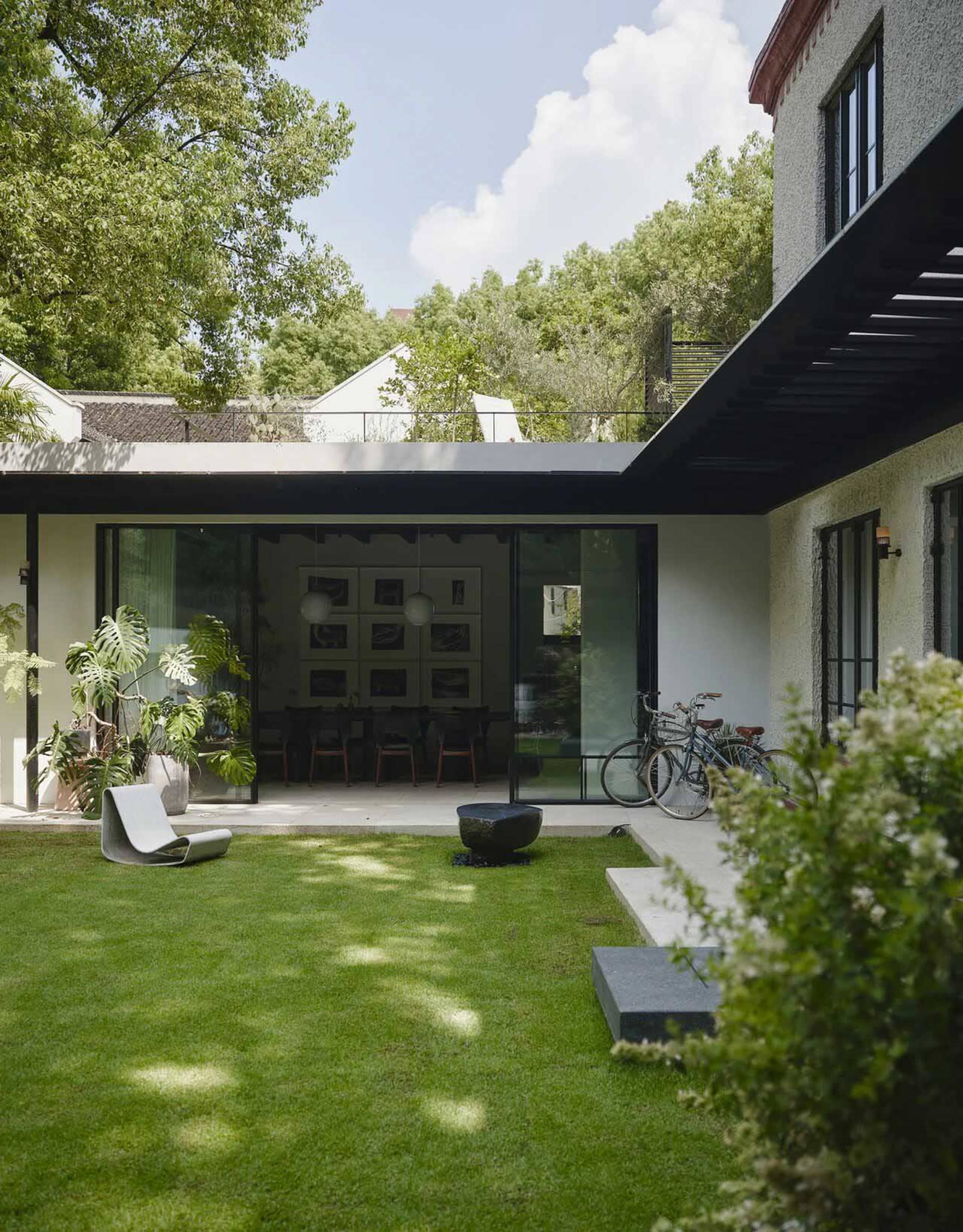 Open living area with a glass door leading to a backyard patio, featuring a dining table, chairs, bicycles, and a kitchen area with a sink and potted plant, bathed in natural light