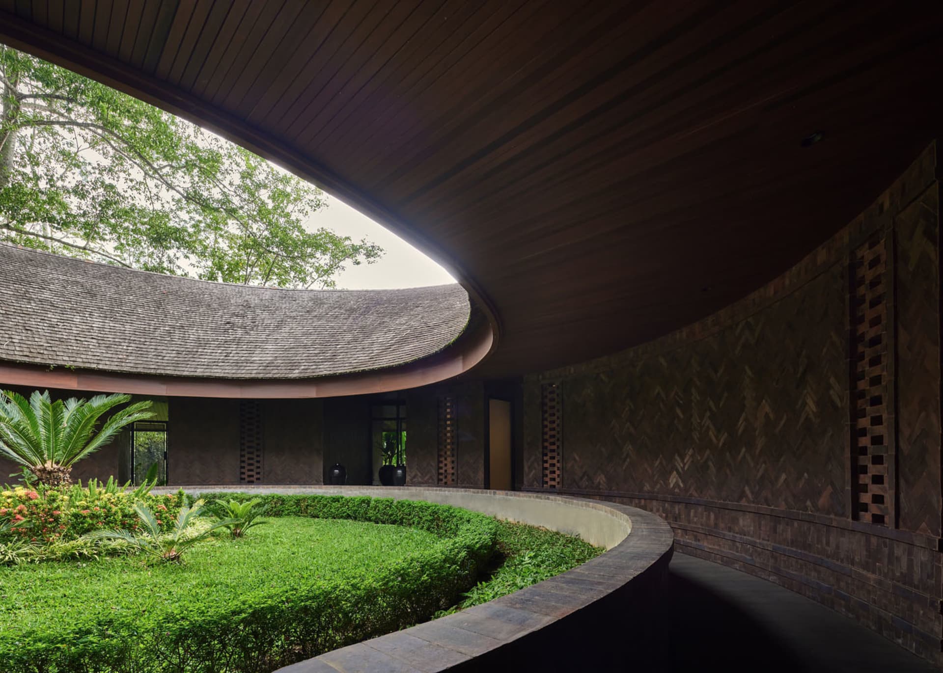 A serene architectural scene featuring a circular building with a wooden dome, surrounded by a lush green garden and potted plants, with two benches for seating