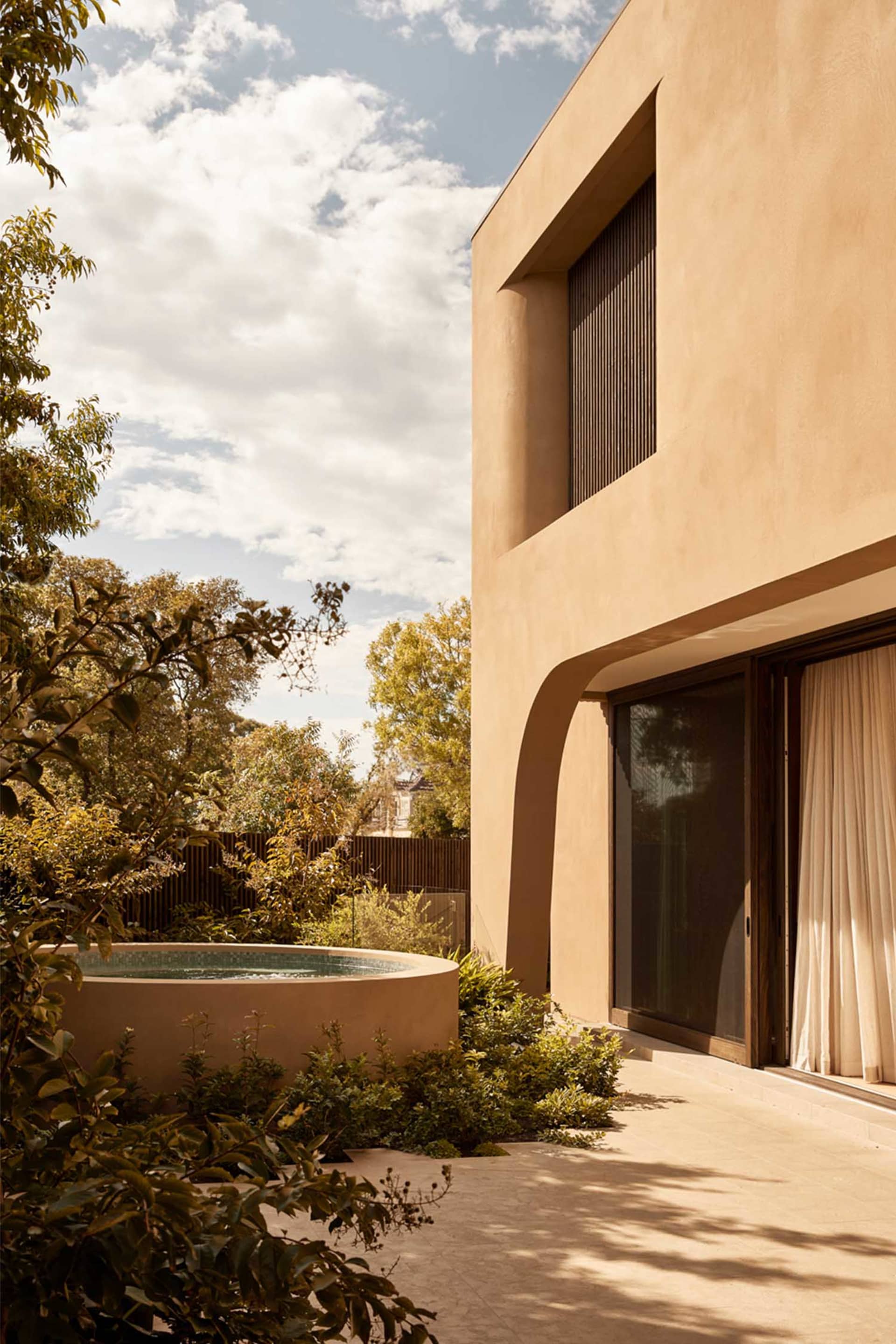A modern house with a large window in front of a tan wall, surrounded by trees and a pool in a warm climate setting