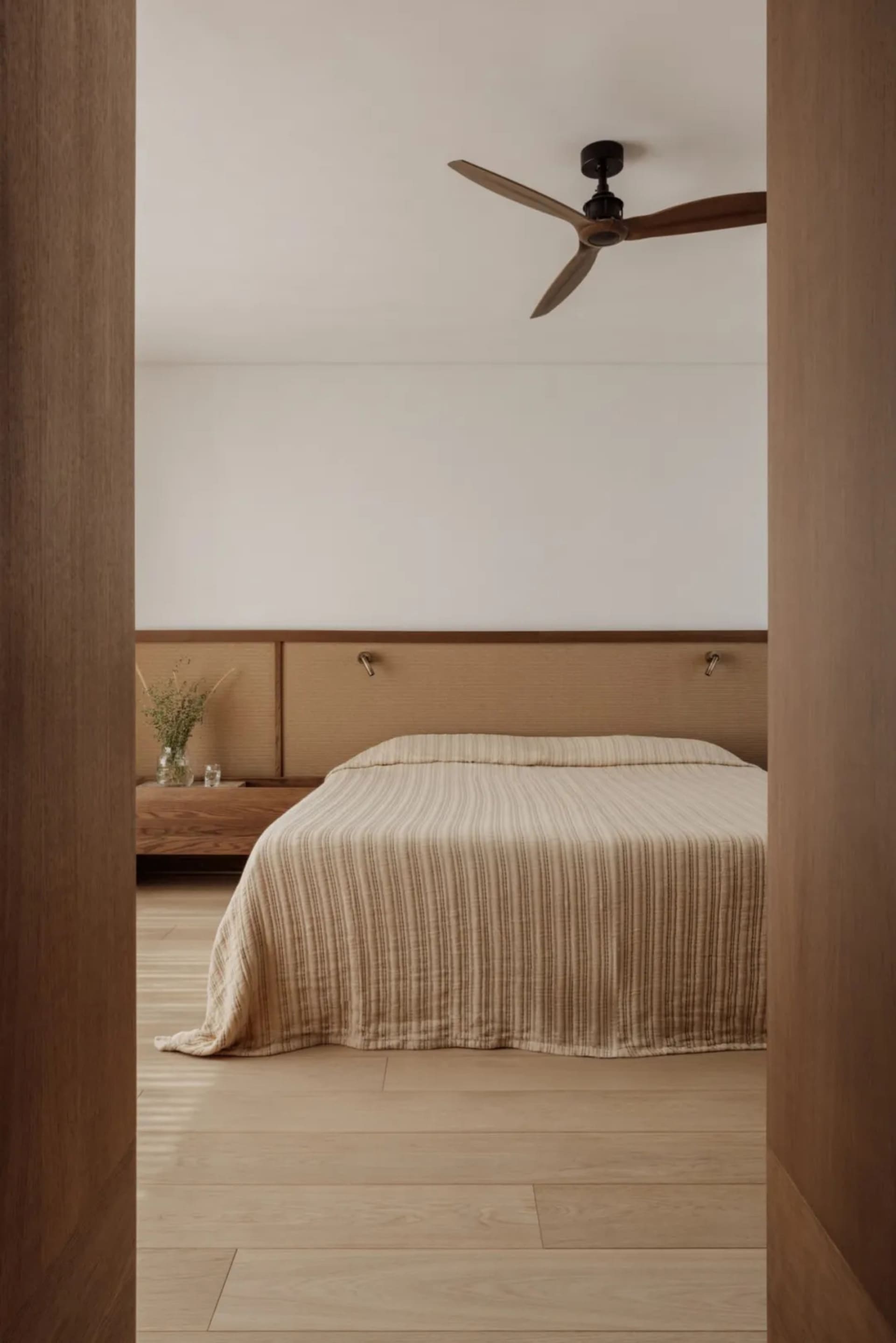 Cozy bedroom with a striped blanket on a hardwood floor, white walls, and a wooden headboard, with two potted plants and a vase nearby