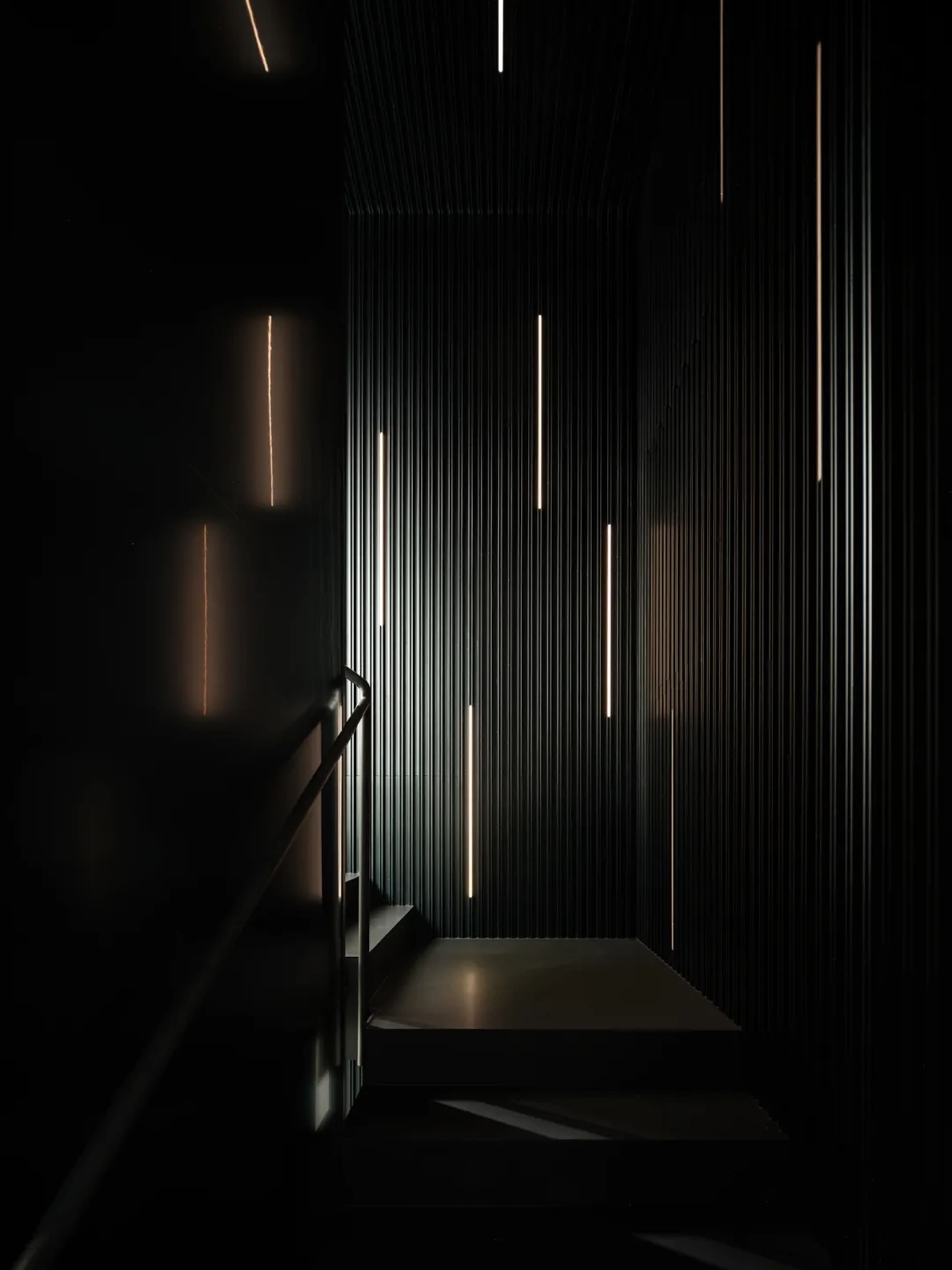 A modern stairwell with a black wall and white railing, illuminated by several lights, leading to a lower level with access to different floors