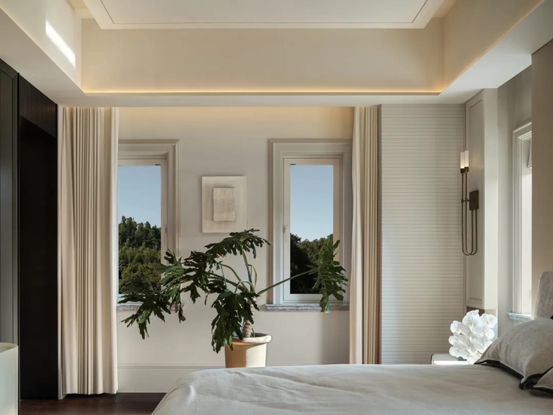 A bright bedroom with a large window, two smaller windows, and a bed extending from one end of the room to the other, with a potted plant near the foot of the bed
