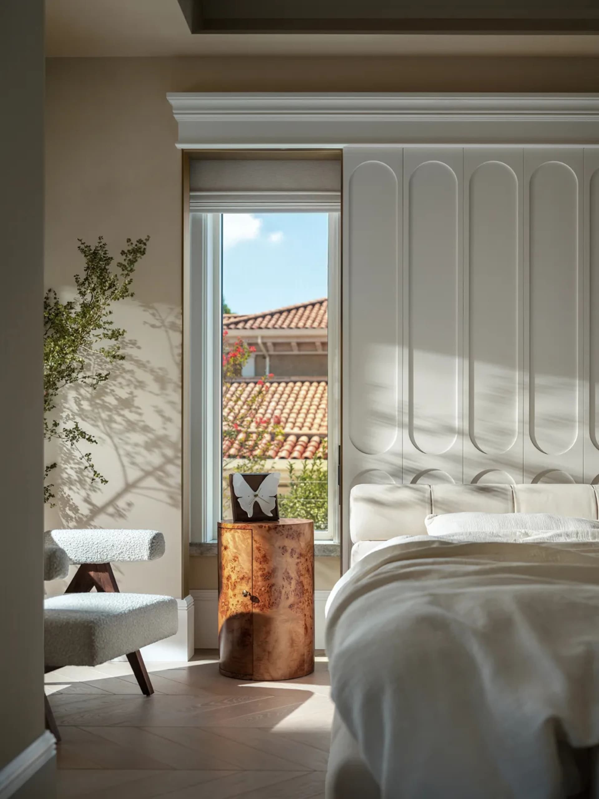 Well-lit bedroom with a white bed, wooden chair, potted plants, and a vase, set against a white wall