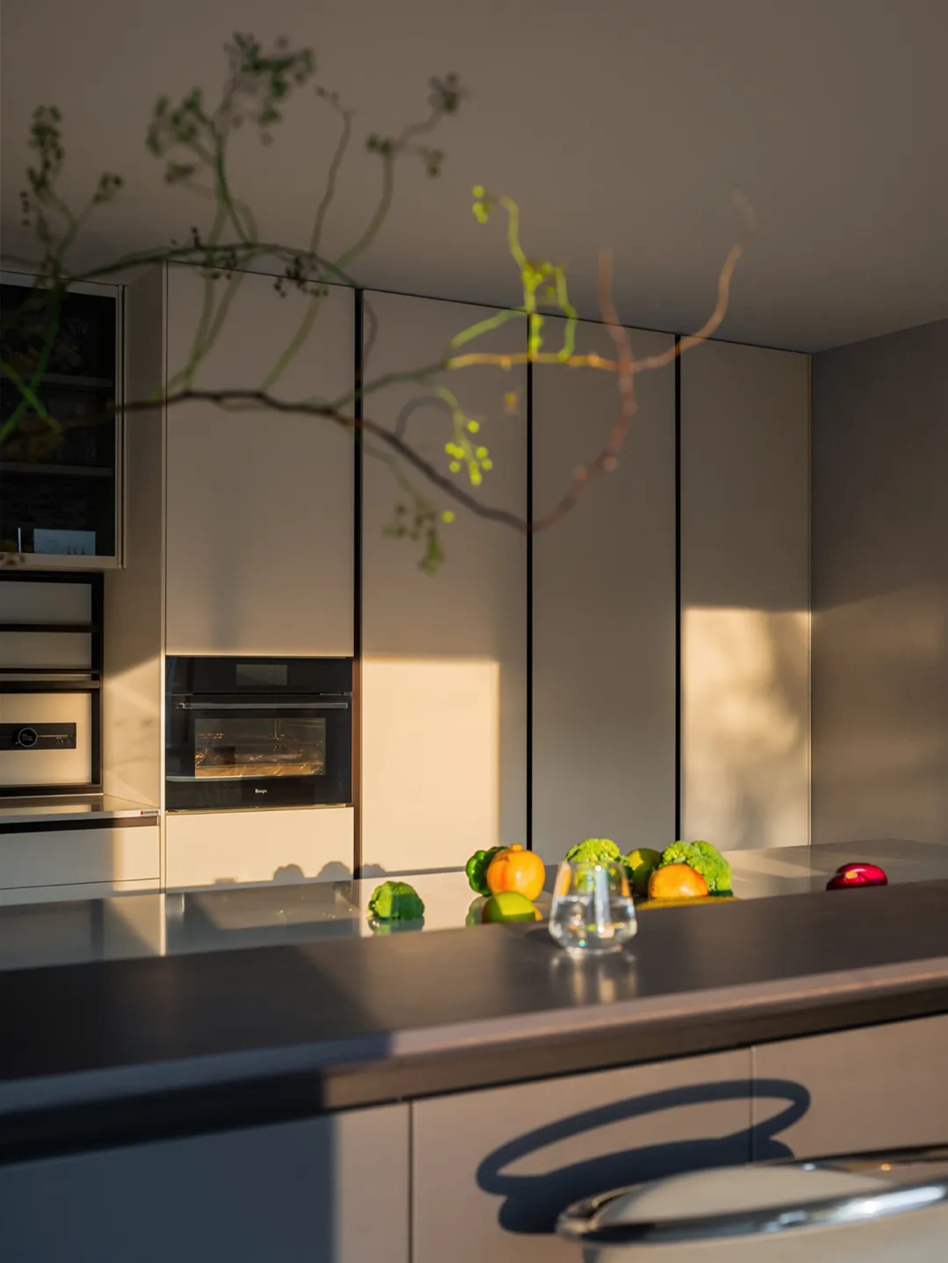 A clean and organized kitchen with white cabinets, a counter with apples and an orange, and a wine glass, set against a neutral background