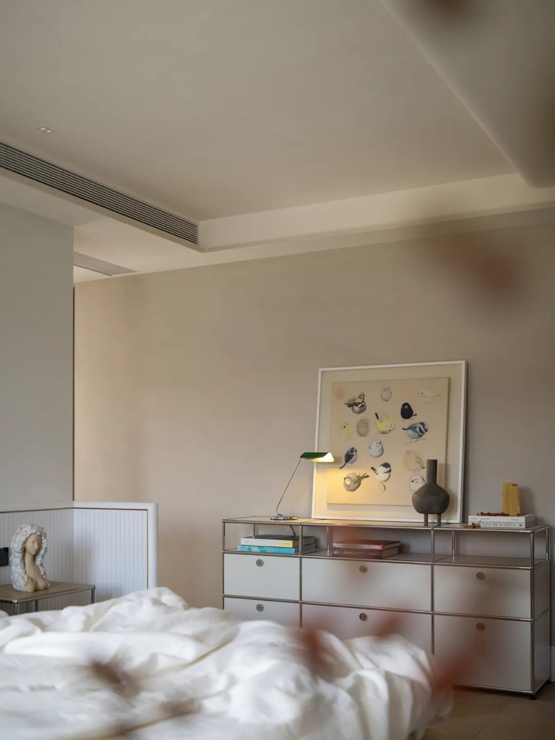 Modern bedroom with a large bed, dresser, nightstand, lamps, and a vase, set in a well-lit space