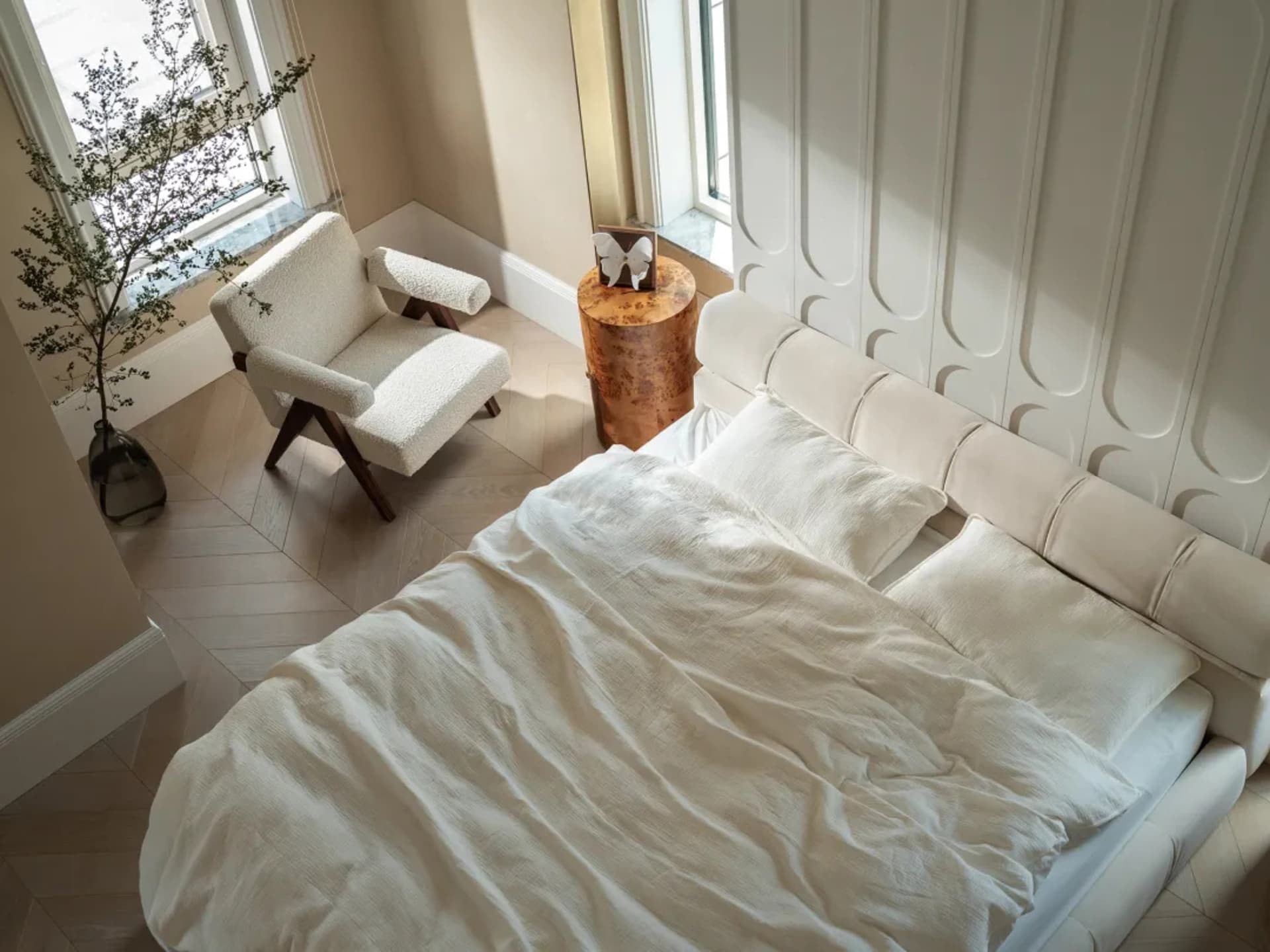 A bedroom with a large white bed on a hardwood floor, featuring two chairs, a potted plant, and a decorative vase, creating a clean and well-organized atmosphere