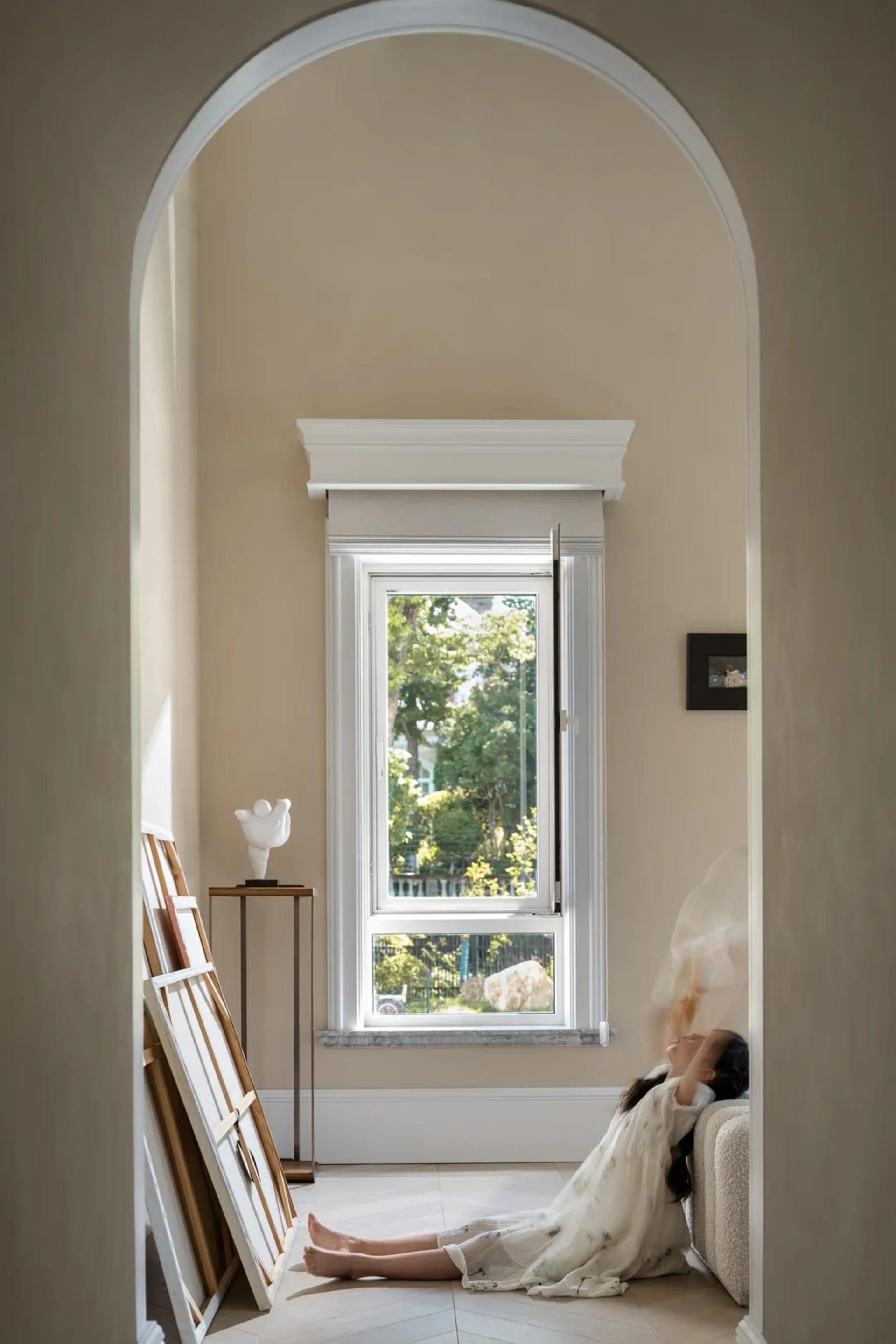A woman sits on a window seat in a room with a white wall, looking out at a car parked outside through a large window, with two vases placed nearby