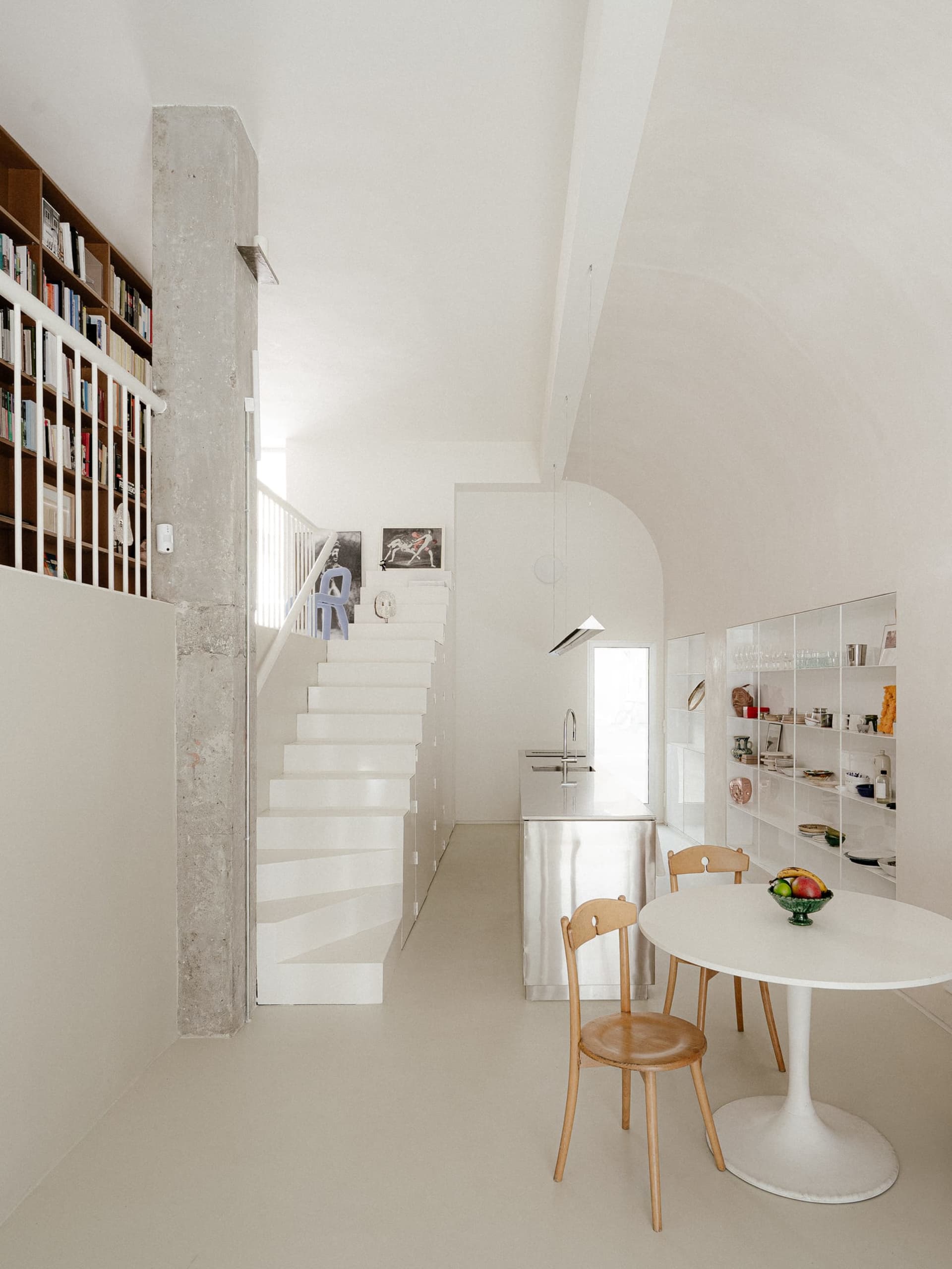 Open living area with a modern kitchen, dining table, and living room, featuring white walls, floors, and stainless steel countertops, with a lived-in atmosphere and multiple books scattered throughout