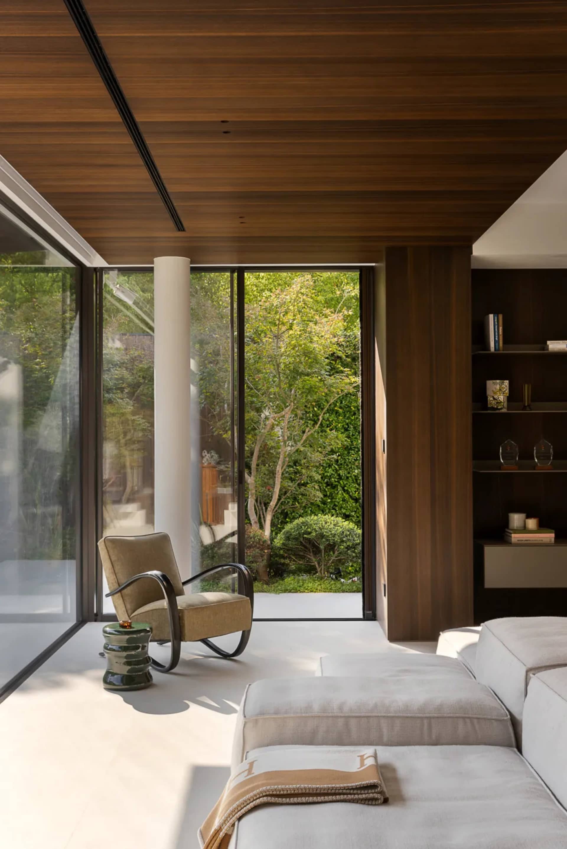 A modern living space with wooden floors and walls, featuring a large window, two couches, chairs, and a bookshelf, with ample natural light and a warm atmosphere