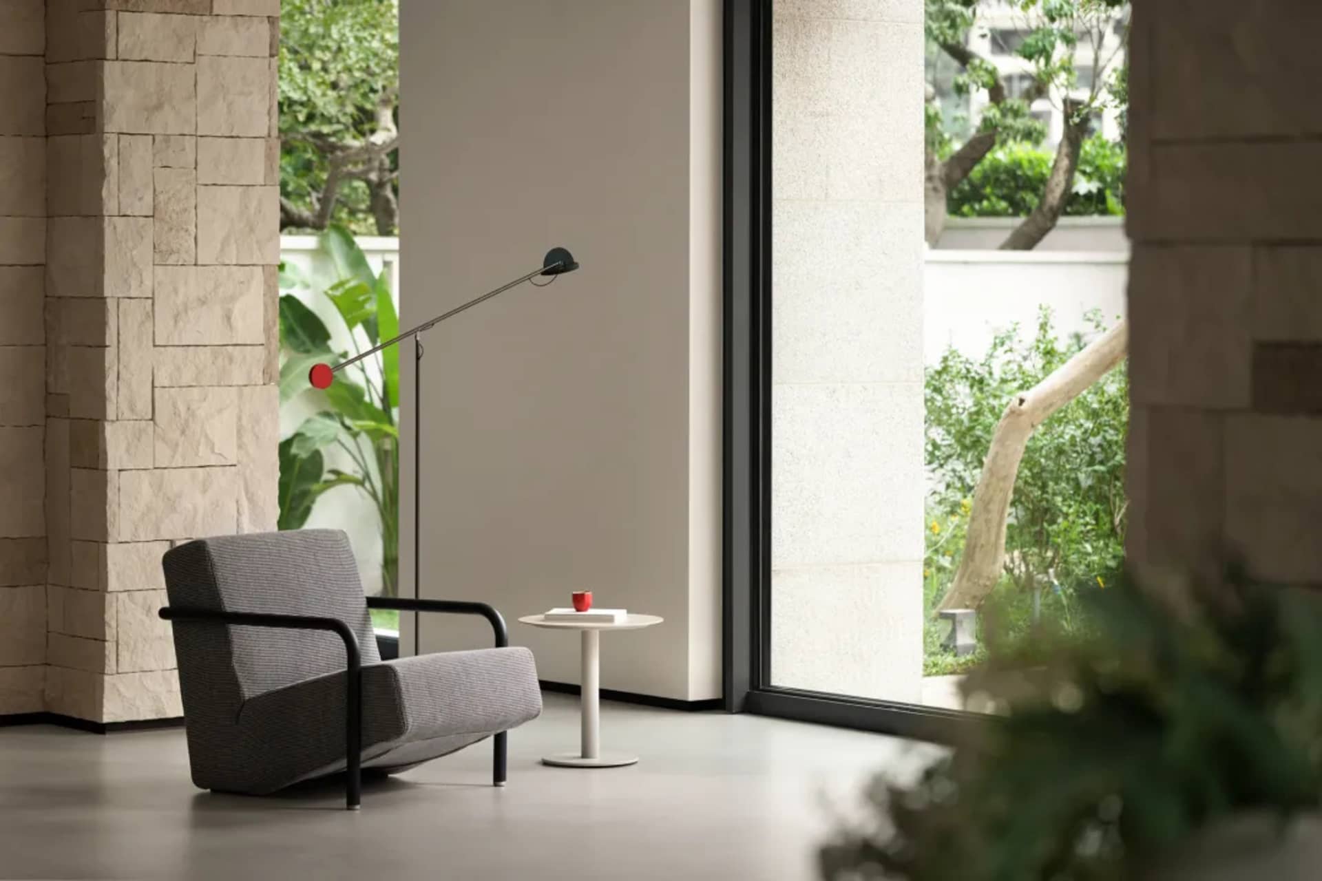 A gray chair sits in front of a window in a calm and inviting living space with natural light and two potted plants nearby